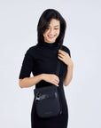 Close up view of a dark haired model smiling down at her bag. She is wearing a black dress and Sherpani's Anti-Theft crossbody the Prima AT in Carbon.