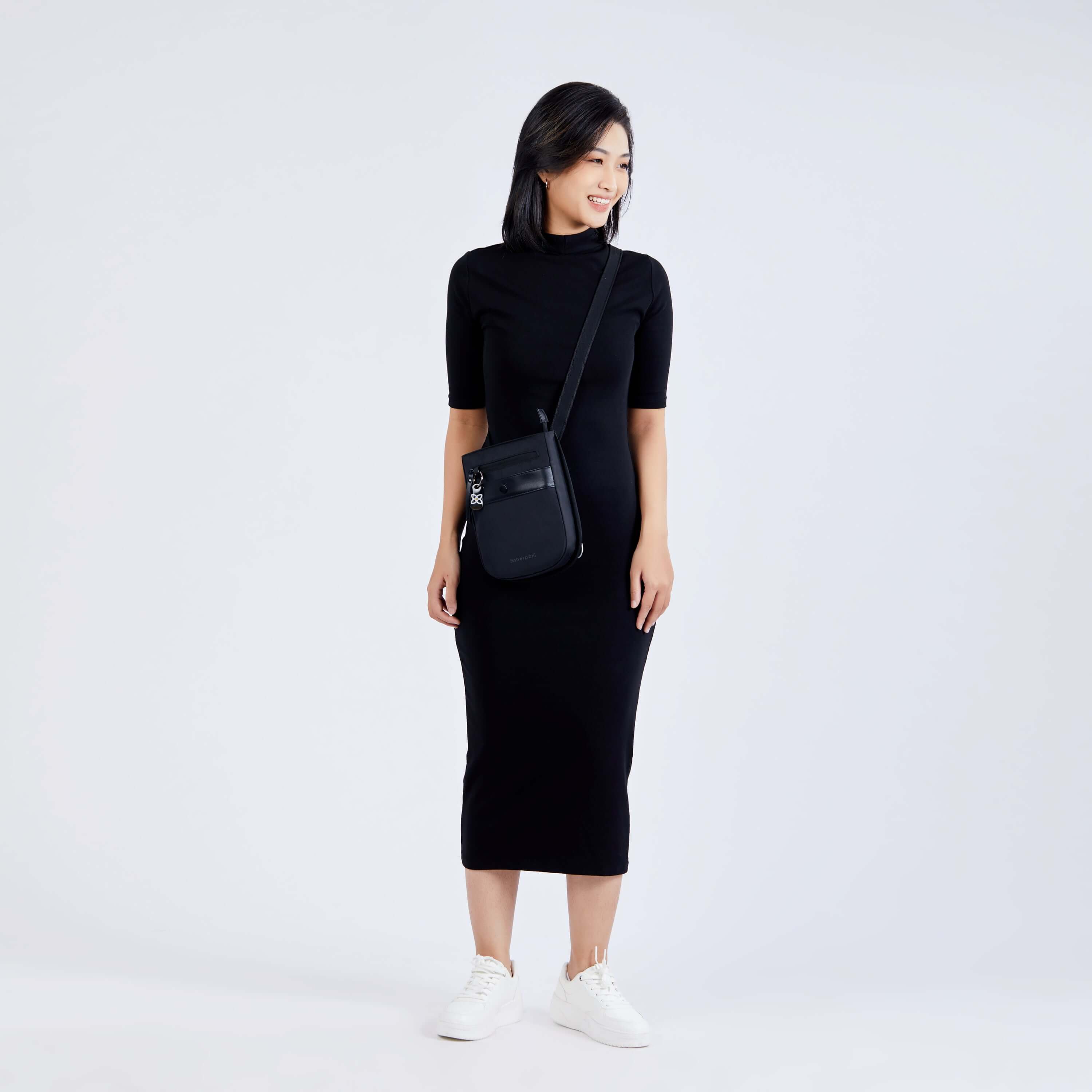 Full body view of a dark haired model facing the camera and smiling over her left shoulder. She is wearing a black dress, white shoes and Sherpani&#39;s Anti-Theft crossbody the Prima AT in Carbon.