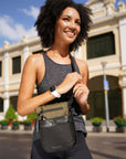 Close up view of curly haired model walking down a city street and smiling. She is wearing a gray tank top, black leggings and Sherpani's Anti-Theft crossbody the Prima AT in Loden.