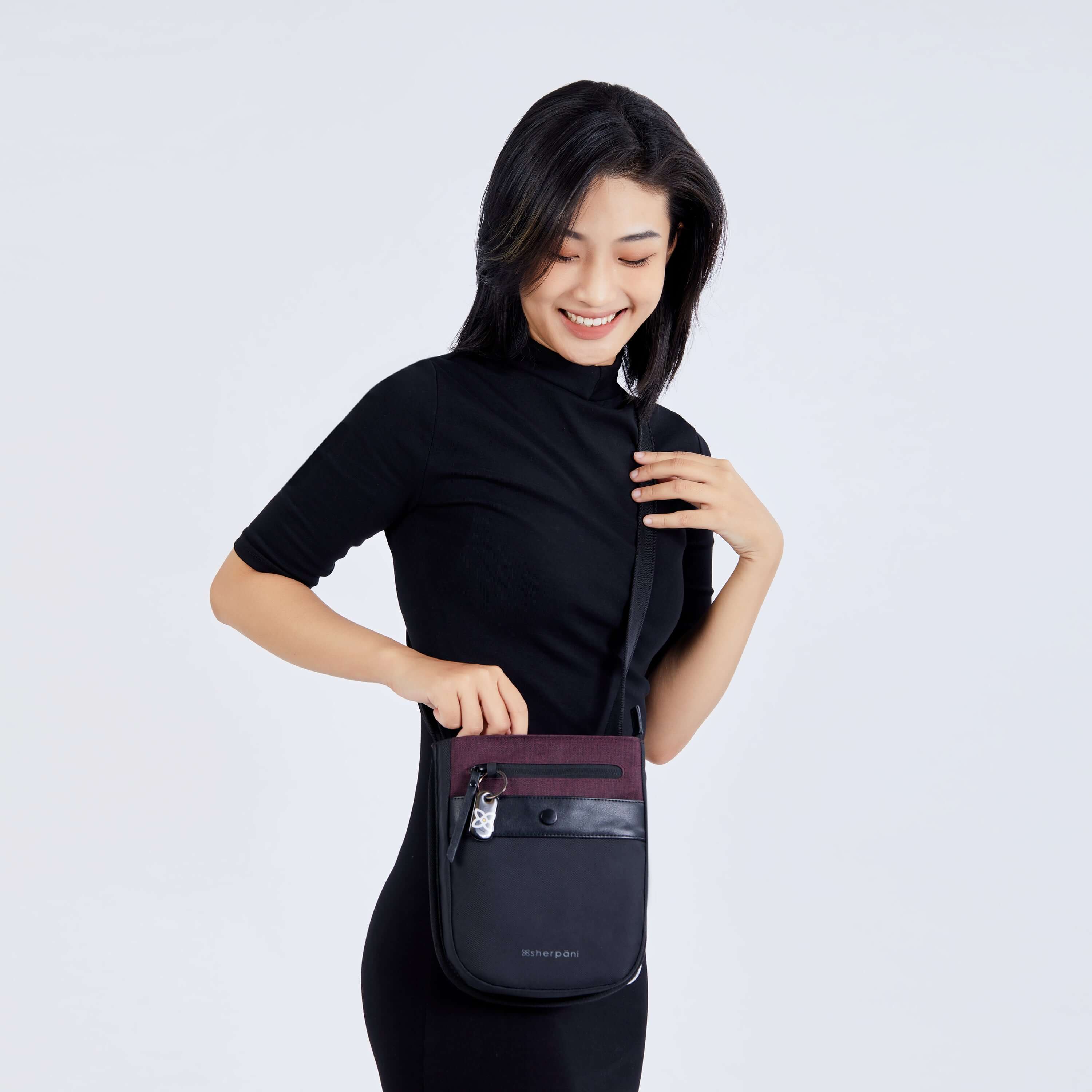 Close up view of a dark haired smiling down at her bag. She is wearing a black dress and Sherpani&#39;s Anti-Theft crossbody the Prima AT in Merlot.