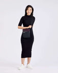 Full body view of a model facing the camera and smiling. She is wearing a black dress, white shoes and Sherpani's Anti-Theft crossbody the Prima AT in Merlot.