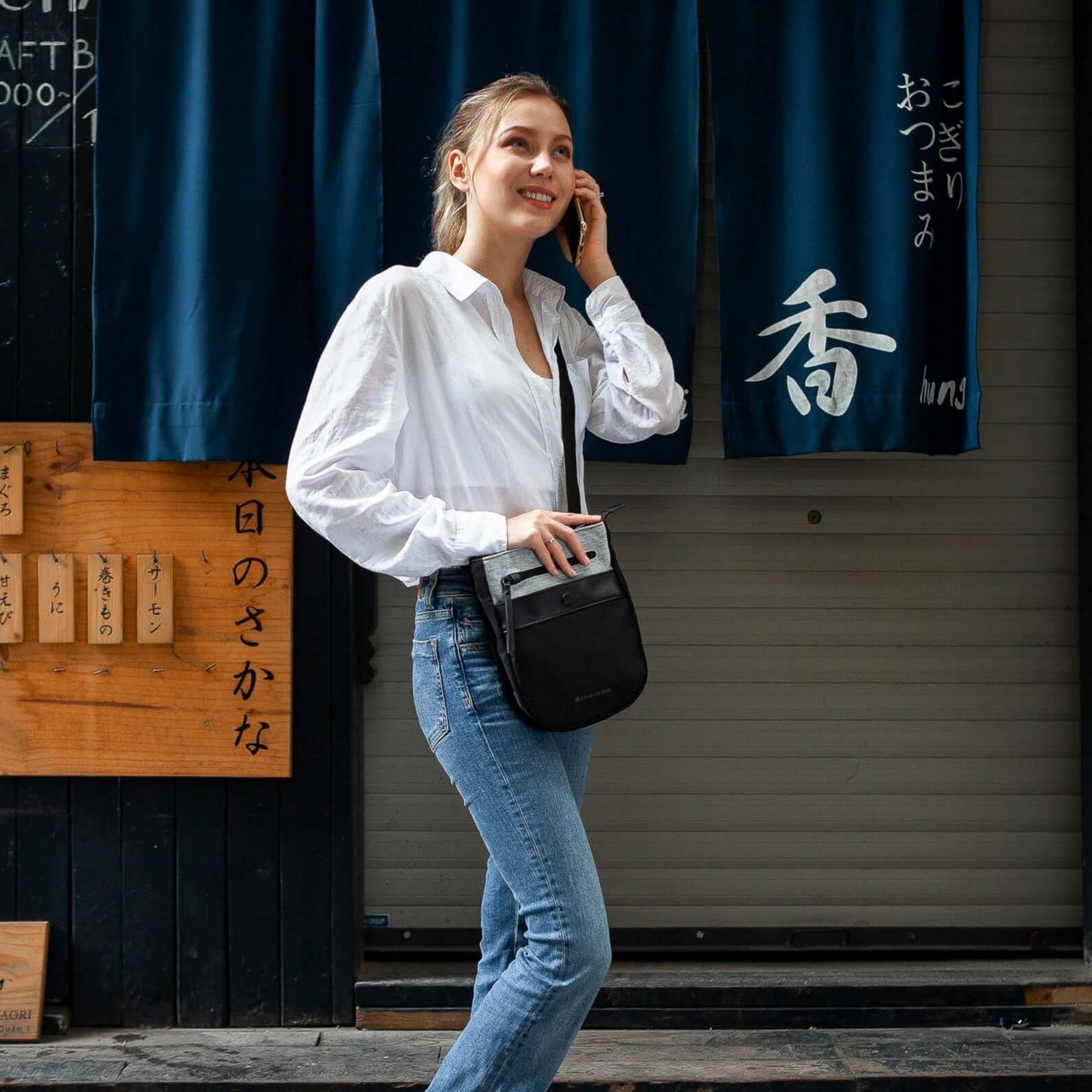Full body view of a model outside of a restaurant talking on the phone. She is wearing a white shirt, blue jeans and Sherpani's Anti-Theft crossbody the Prima AT in Sterling. 