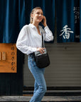 Full body view of a model outside of a restaurant talking on the phone. She is wearing a white shirt, blue jeans and Sherpani's Anti-Theft crossbody the Prima AT in Sterling.