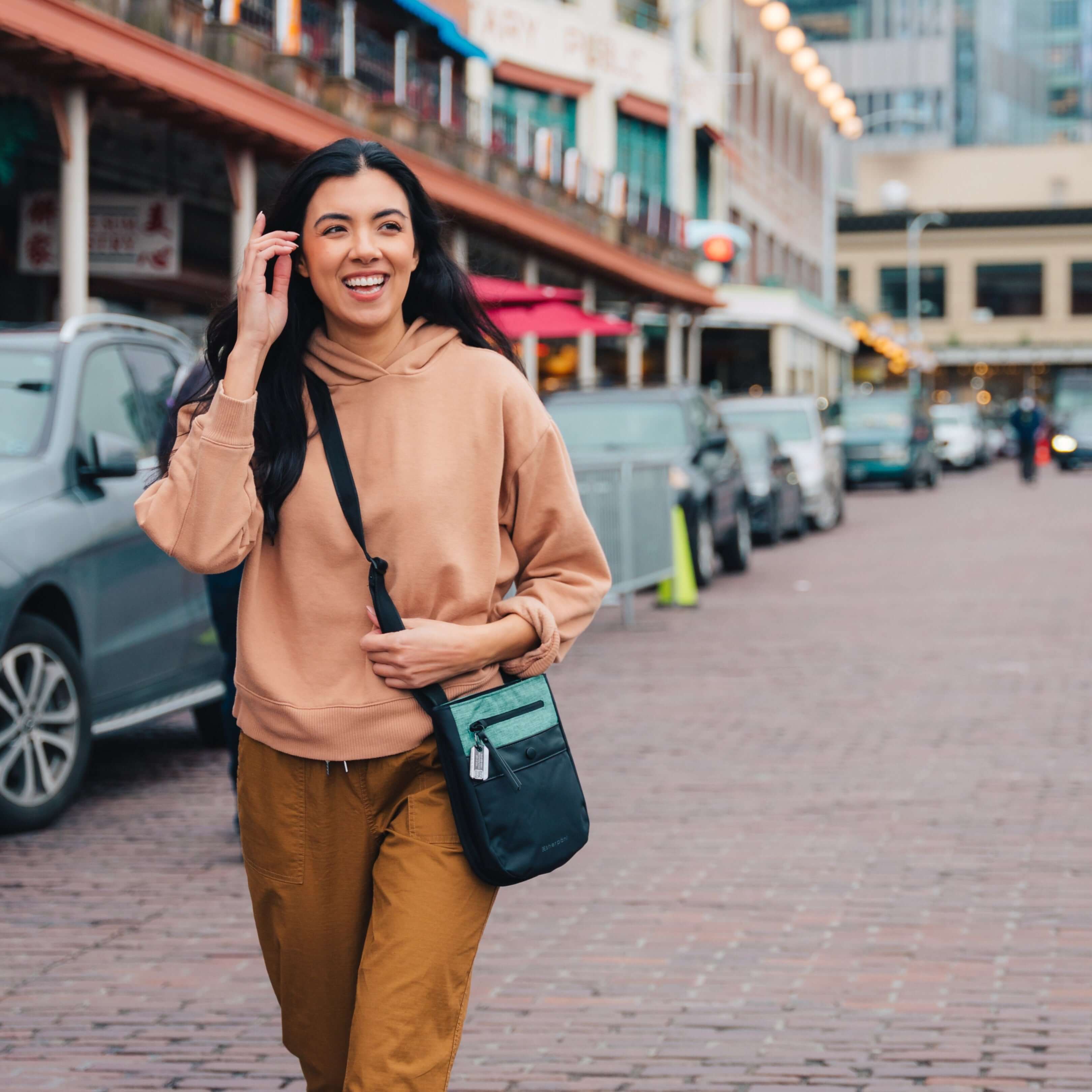 A dark haired model is walking on the street. She is wearing a sweater, orange pants and Sherpani&#39;s Anti-Theft crossbody the Prima AT in Teal.