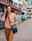 A dark haired model is walking on the street. She is wearing a sweater, orange pants and Sherpani's Anti-Theft crossbody the Prima AT in Teal.