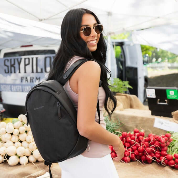 A dark haired woman stands outside in a farmer's market, she smiles over her right shoulder at the camera. She is wearing a sunglasses, a pink tank top, white leggings and Sherpani's Anti-Theft backpack, the Presta in Black. 