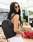 A dark haired woman stands outside in a farmer's market, she smiles over her right shoulder at the camera. She is wearing a sunglasses, a pink tank top, white leggings and Sherpani's Anti-Theft backpack, the Presta in Black.