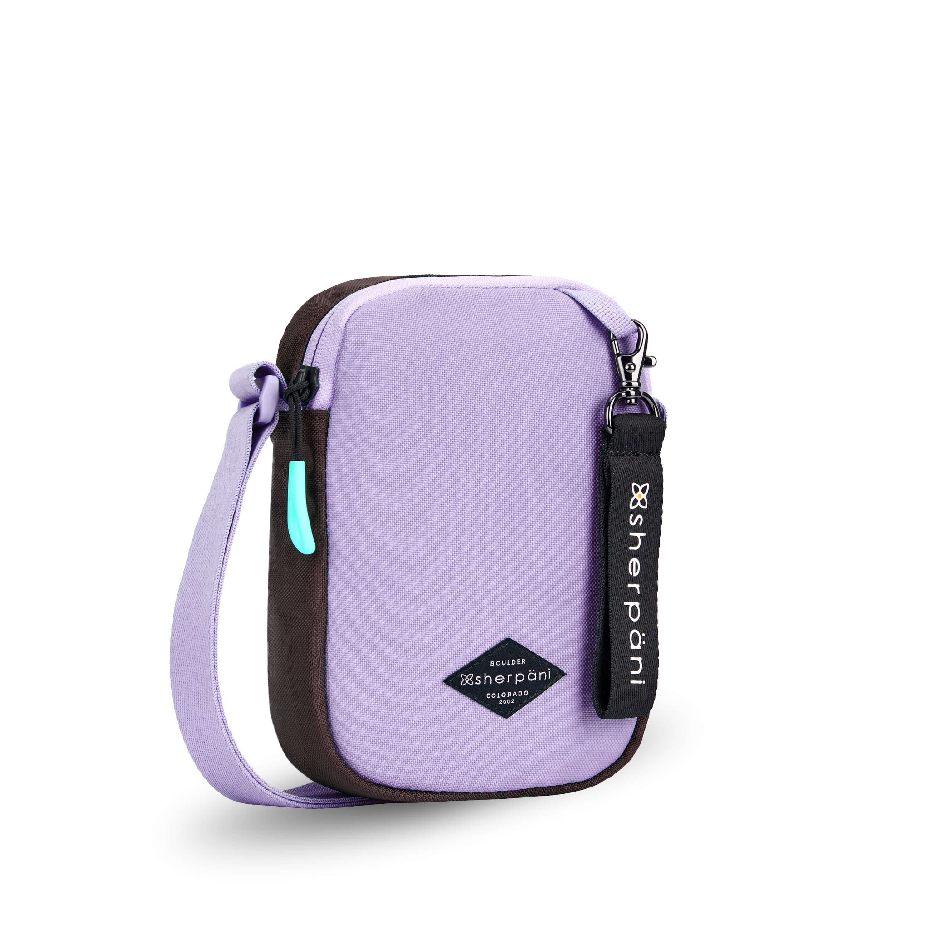 Angled front view of Sherpani crossbody, the Rogue in Lavender. The bag is two toned: the front is lavender and the back is brown. The main zipper compartment features an easy-pull zipper accented in aqua. The bag has an adjustable crossbody strap. A branded Sherpani keychain is clipped to a fabric loop on the top right corner. #color_lavender