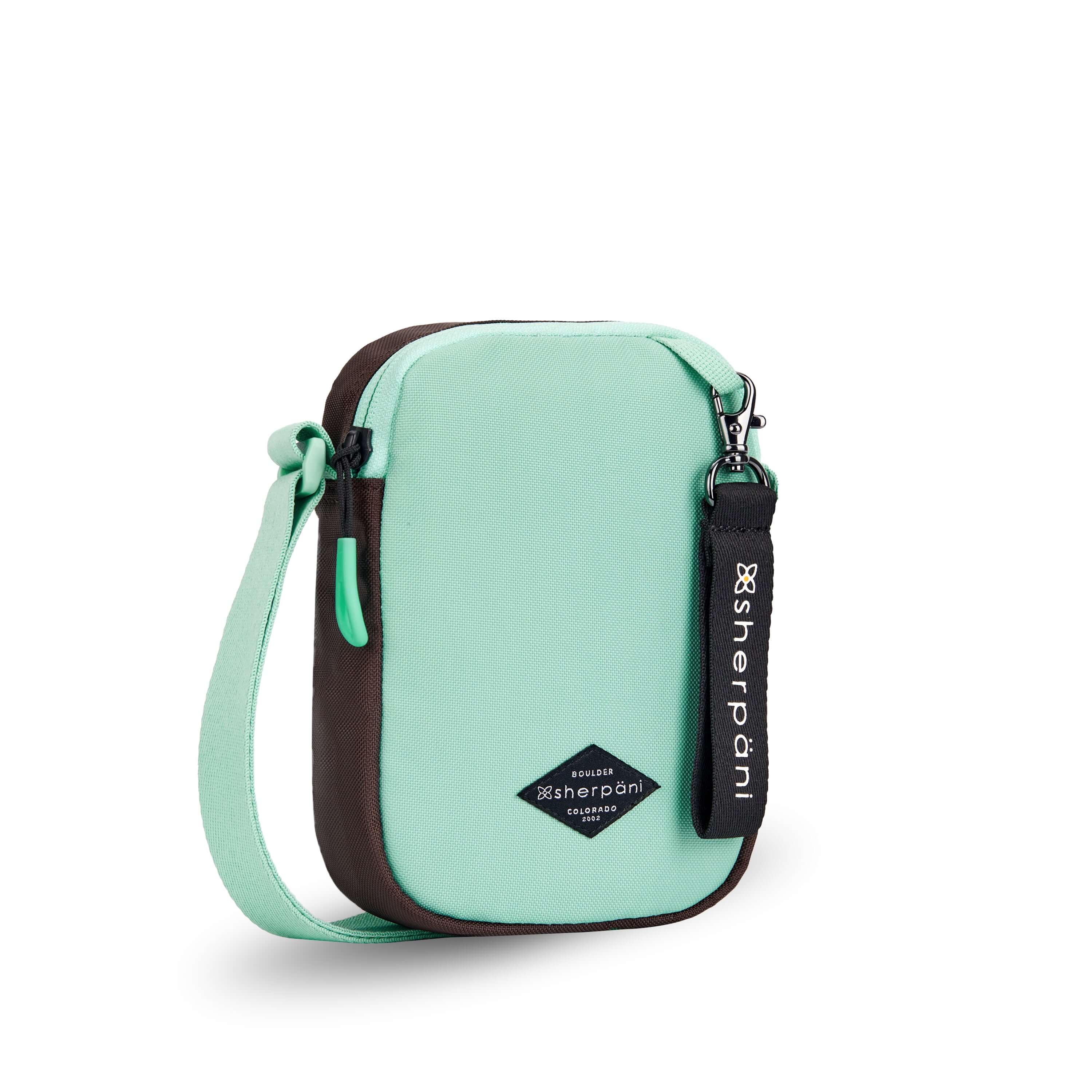 Angled front view of Sherpani crossbody, the Rogue in Seagreen. The bag is two toned: the front is light green and the back is brown. The main zipper compartment features an easy-pull zipper accented in light green. The bag has an adjustable crossbody strap. A branded Sherpani keychain is clipped to a fabric loop on the top right corner. #color_seagreen