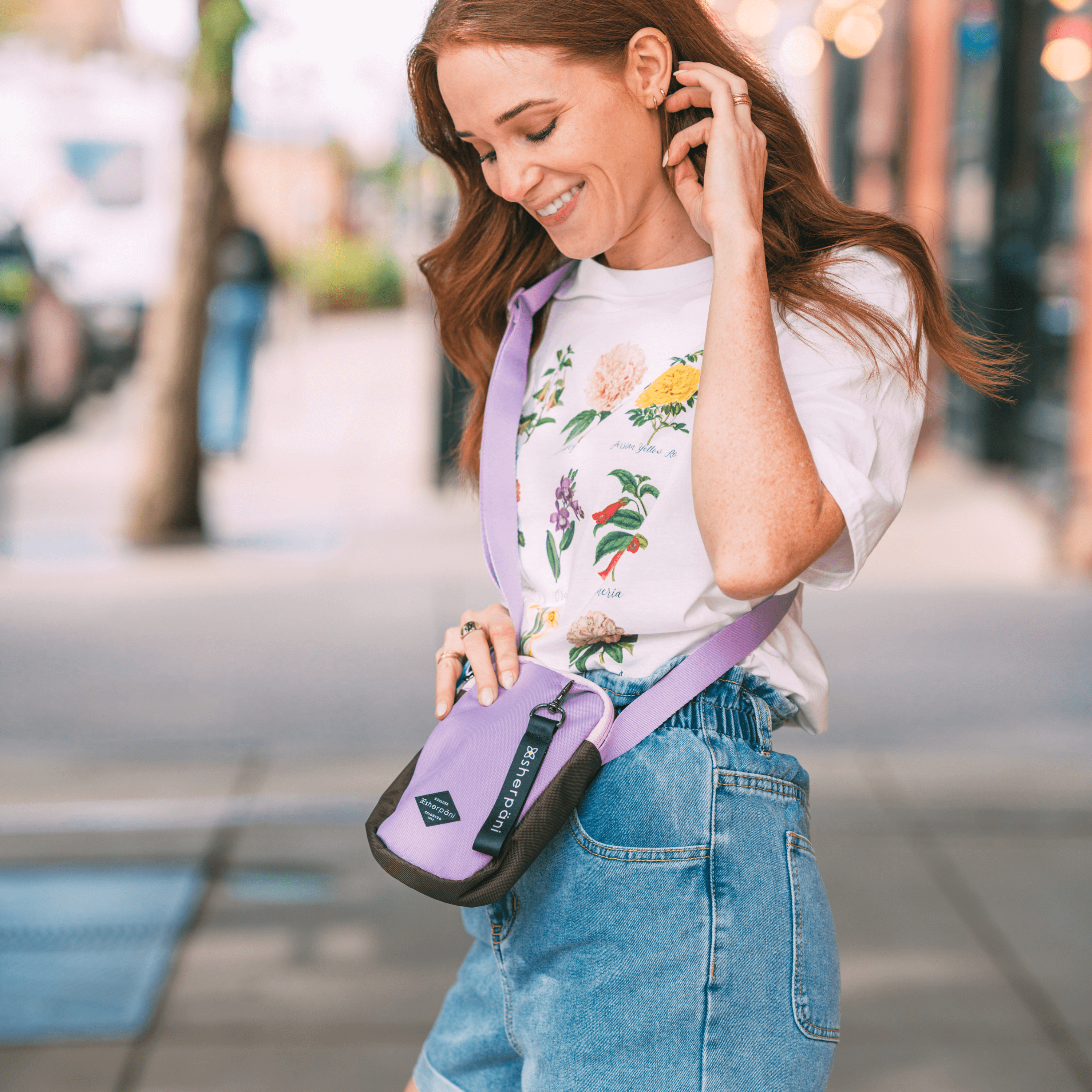 A red haired woman stands outside on the sidewalk. She is wearing a white shirt with flowers and jeans. She is smiling downward at the Sherpani crossbody, the Rogue in Lavender.