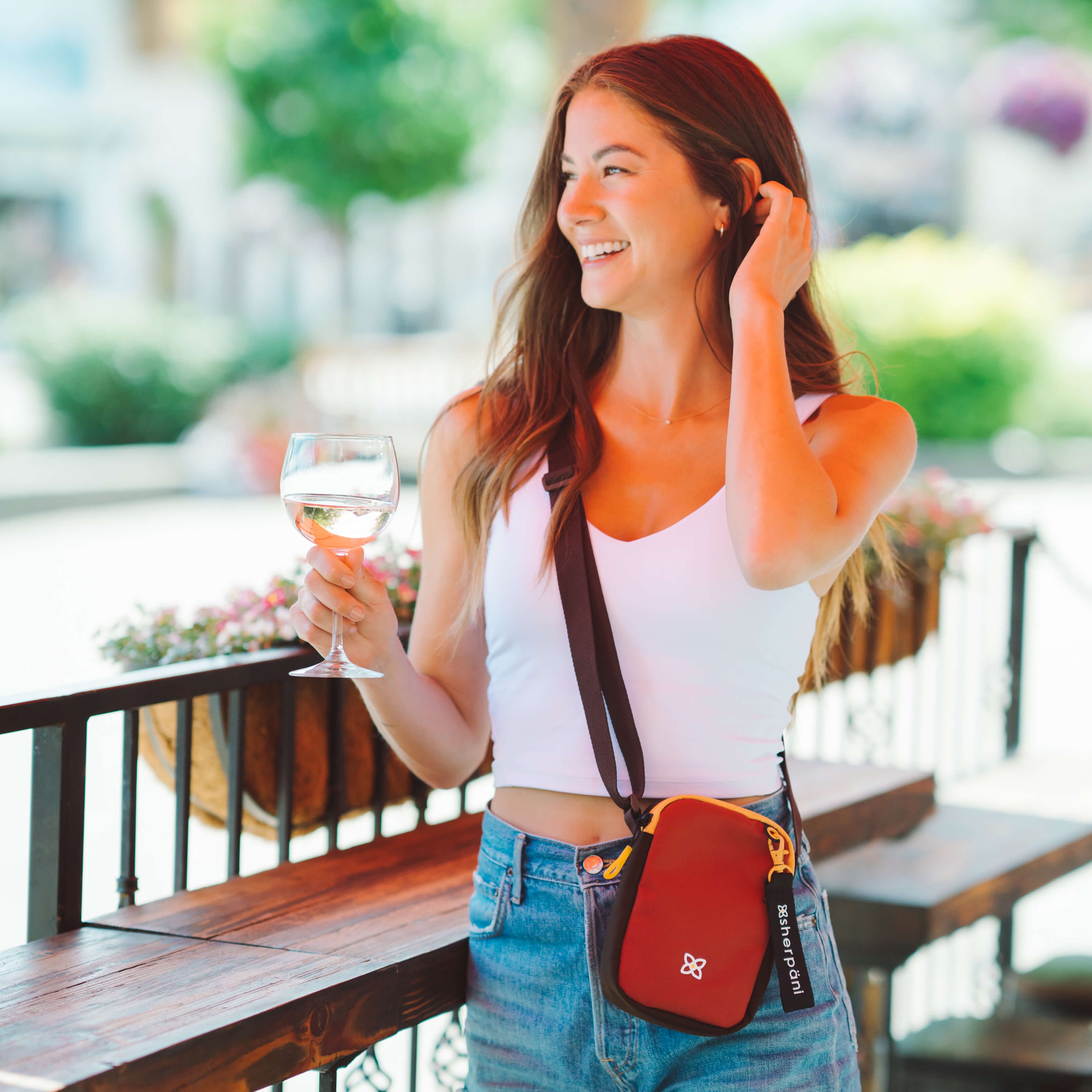 A woman standing on an outdoor patio holds up a glass of wine. She is wearing Sherpani RFID crossbody purse, the Rogue in Cider. 