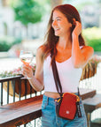 A woman standing on an outdoor patio holds up a glass of wine. She is wearing Sherpani RFID crossbody purse, the Rogue in Cider.