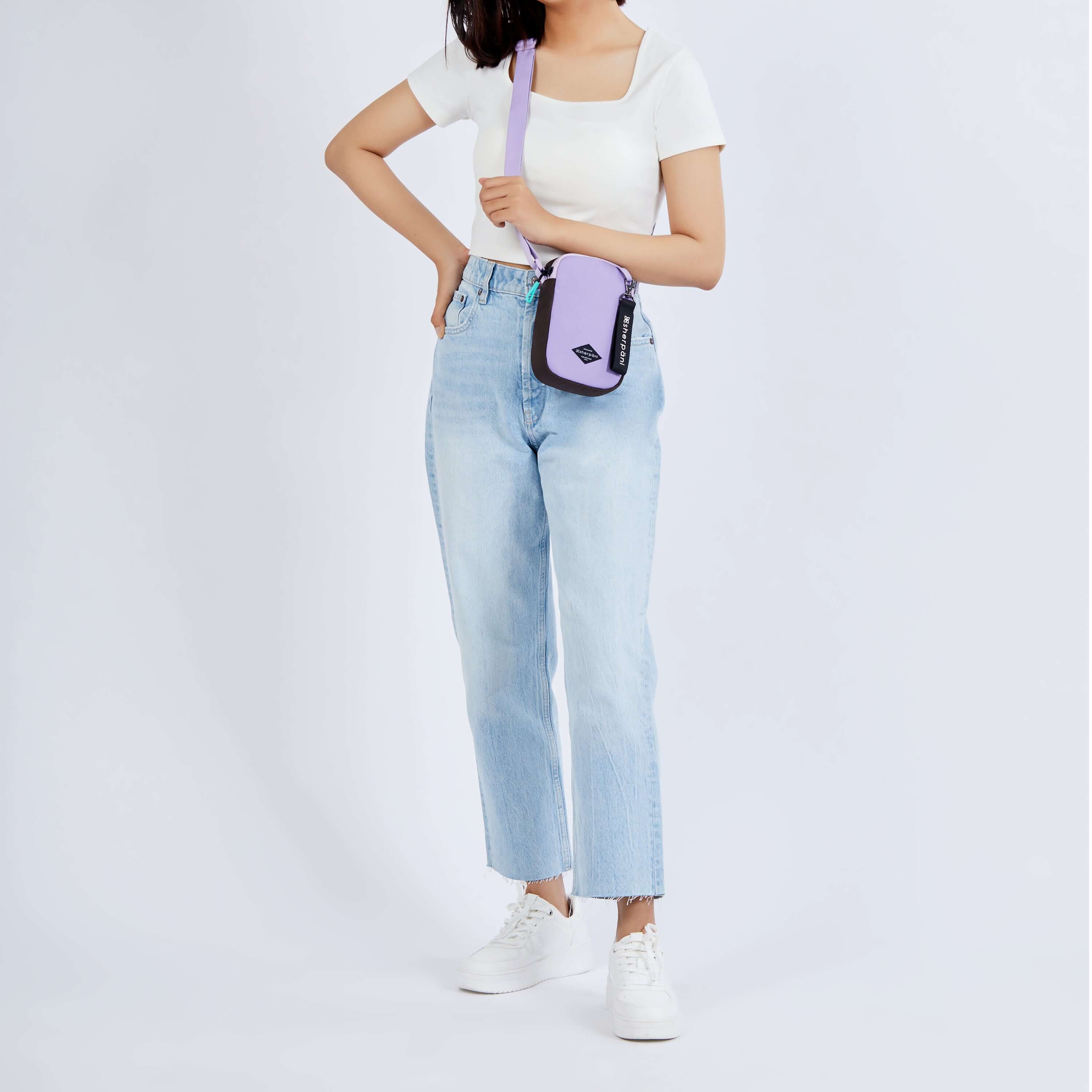 Full body view of a dark haired model facing the camera. She is wearing a white tee shirt, jeans and white sneakers. She carries Sherpani crossbody, the Rogue in Lavender, as a crossbody.