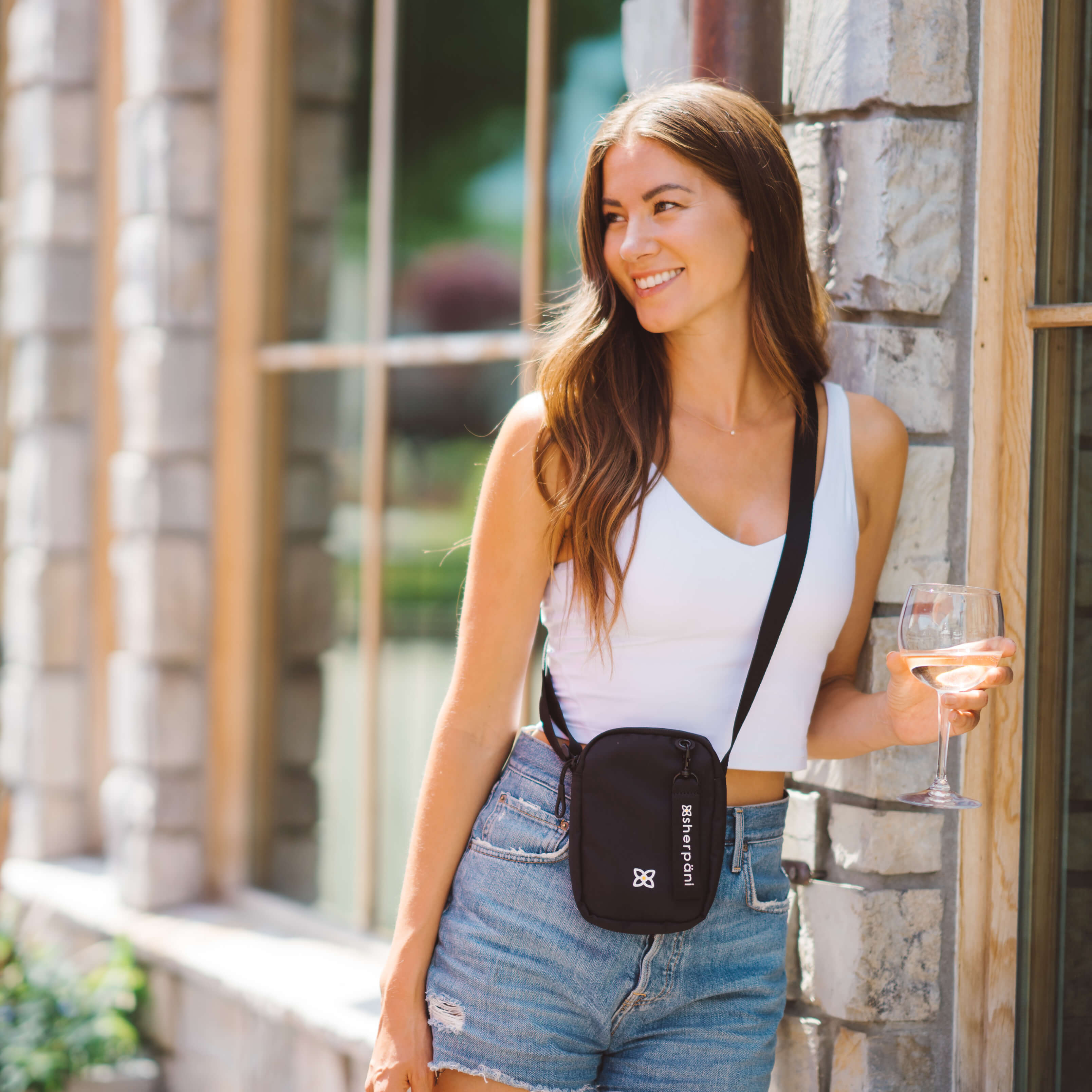 A woman leans against a wall outside with a glass of wine in her had. She is wearing Sherpani RFID blocking crossbody bag, the Rogue in Raven.