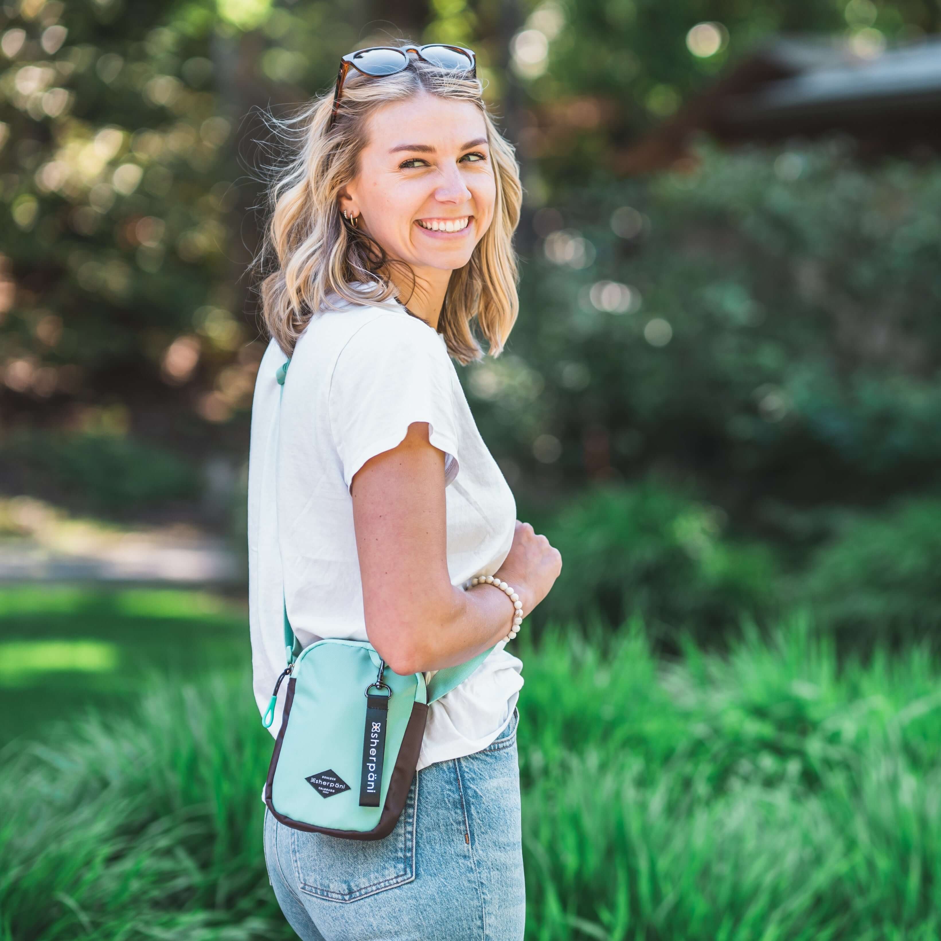 A blonde woman stands outside in a park. She is facing the side and smiling over her right shoulder. She is wearing a white tee shirt and jeans. She carries Sherpani crossbody, the Rogue in Seagreen, as a crossbody.