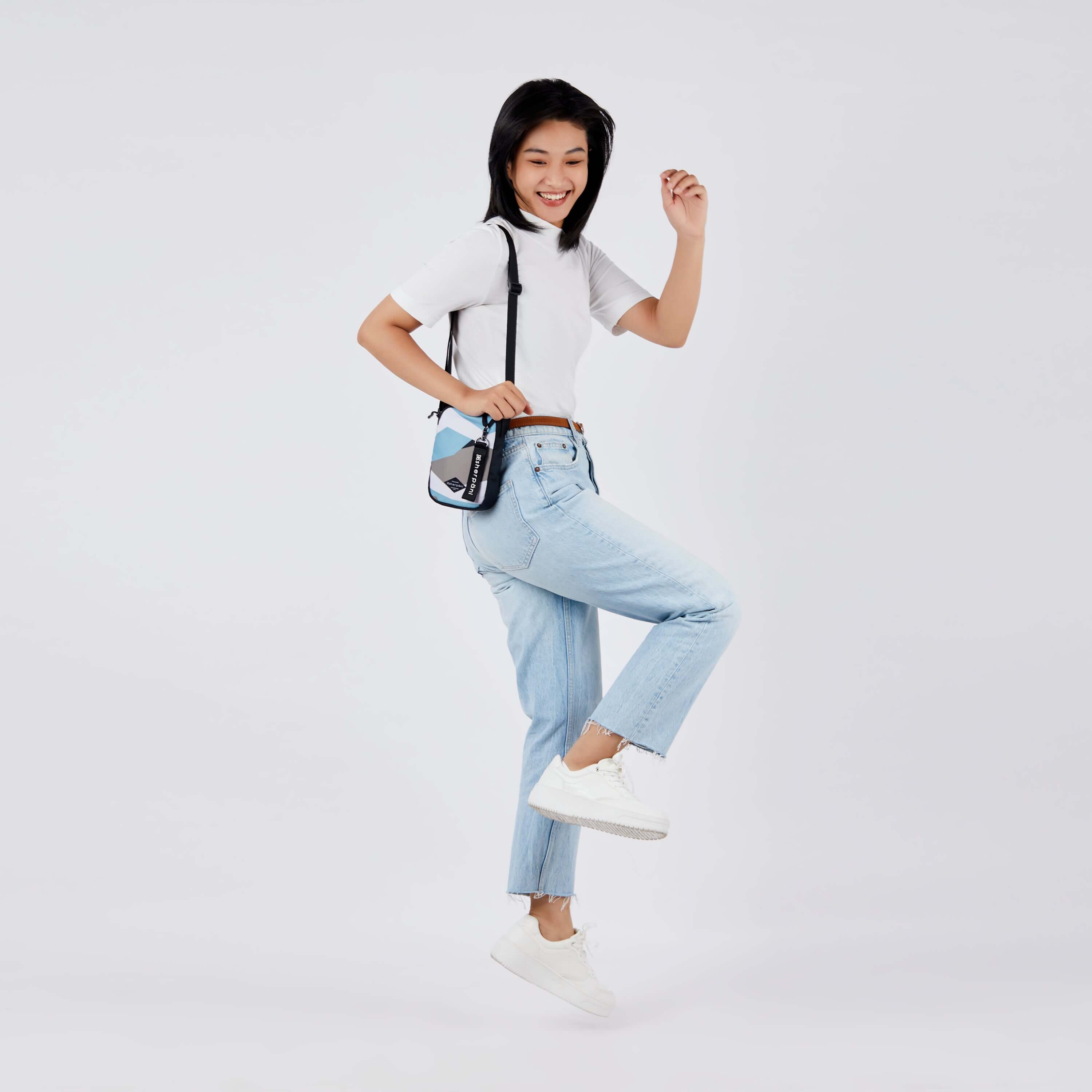 Full body view of a dark hard model facing the side and skipping. She is wearing a white tee shirt, jeans and white sneakers. She carries Sherpani crossbody, the Rogue in Summer Camo, over her shoulder.