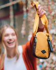 Close up photo of a blonde woman outside in front of an artsy wall. She is holding up Sherpani crossbody, the Rogue in Sundial, by its strap.