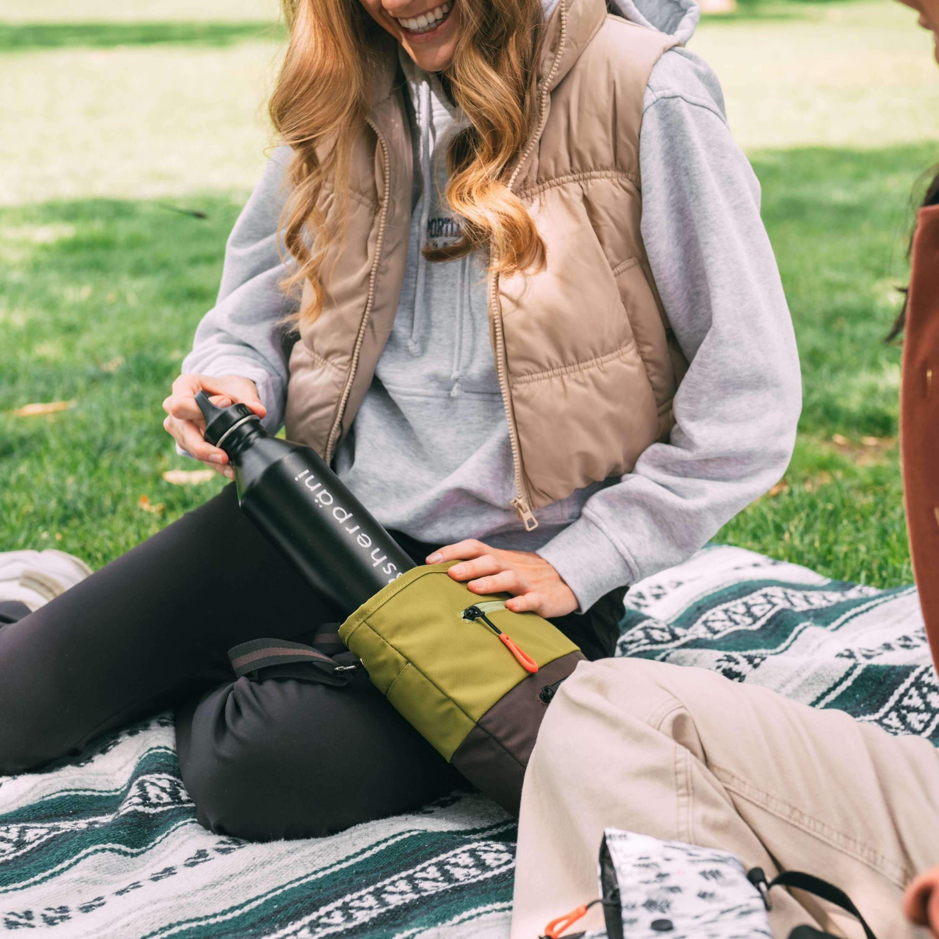 A blonde woman smiles and sits on a picnic blanket outside in the grass. She is wearing a tan vest, gray sweatshirt and black pants. She is pulling a water bottle out of Sherpani crossbody, the Sadie in Cactus. 