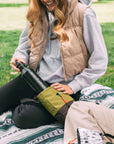 A blonde woman smiles and sits on a picnic blanket outside in the grass. She is wearing a tan vest, gray sweatshirt and black pants. She is pulling a water bottle out of Sherpani crossbody, the Sadie in Cactus.
