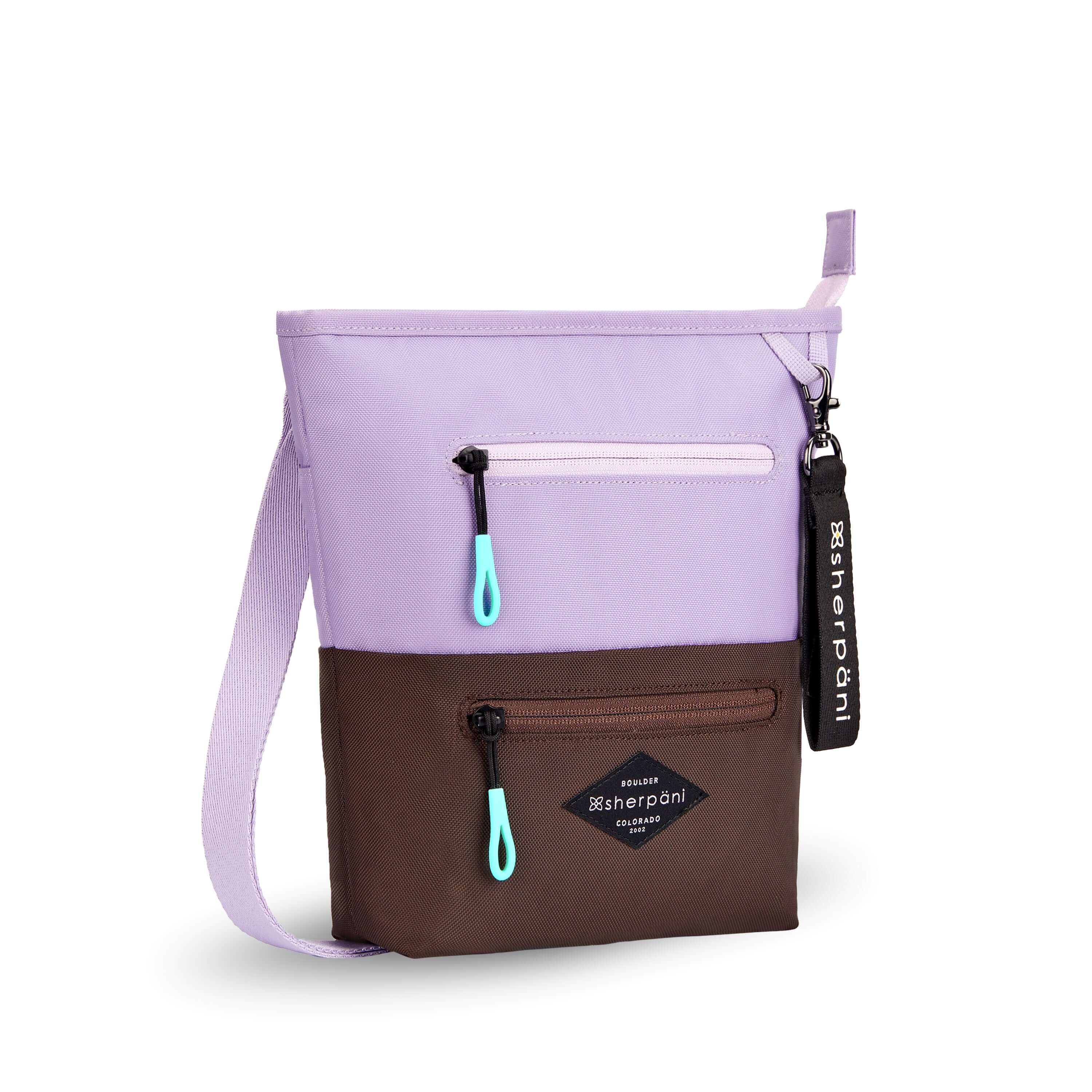 Angled front view of Sherpani’s crossbody, the Sadie, in Lavender. The top half of the bag is lavender and the bottom half of the bag is brown. It features two exterior zipper pockets on the front panel with easy-pull zippers accented in aqua. A Sherpani branded keychain is attached to a fabric loop in the upper right corner. It has an adjustable crossbody strap.