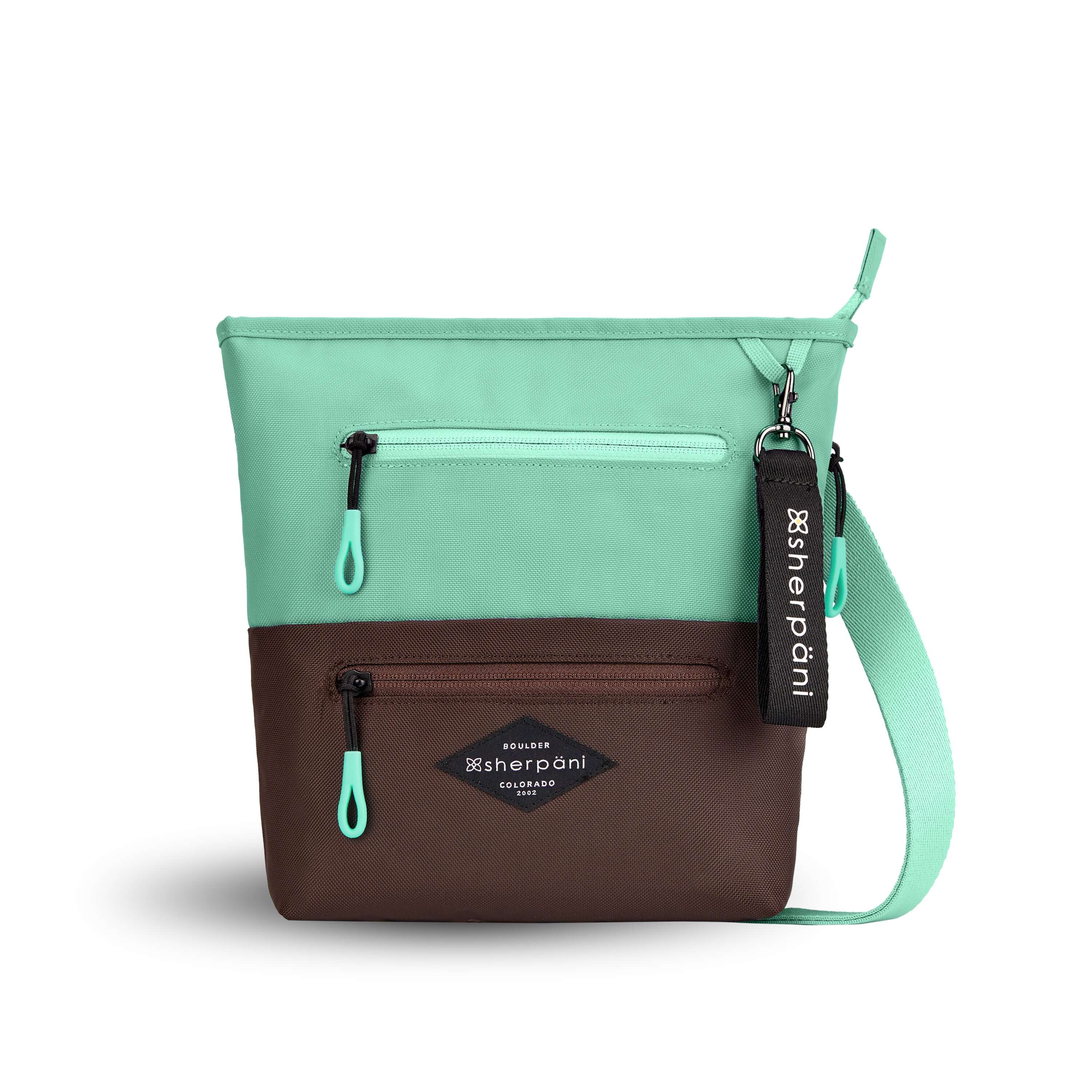 Flat front view of Sherpani’s crossbody, the Sadie, in Seagreen. The top half of the bag is light green and the bottom half of the bag is brown. It features two exterior zipper pockets on the front panel with easy-pull zippers accented in light green. A Sherpani branded keychain is attached to a fabric loop in the upper right corner. It has an adjustable crossbody strap. 