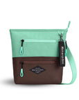Flat front view of Sherpani’s crossbody, the Sadie, in Seagreen. The top half of the bag is light green and the bottom half of the bag is brown. It features two exterior zipper pockets on the front panel with easy-pull zippers accented in light green. A Sherpani branded keychain is attached to a fabric loop in the upper right corner. It has an adjustable crossbody strap.