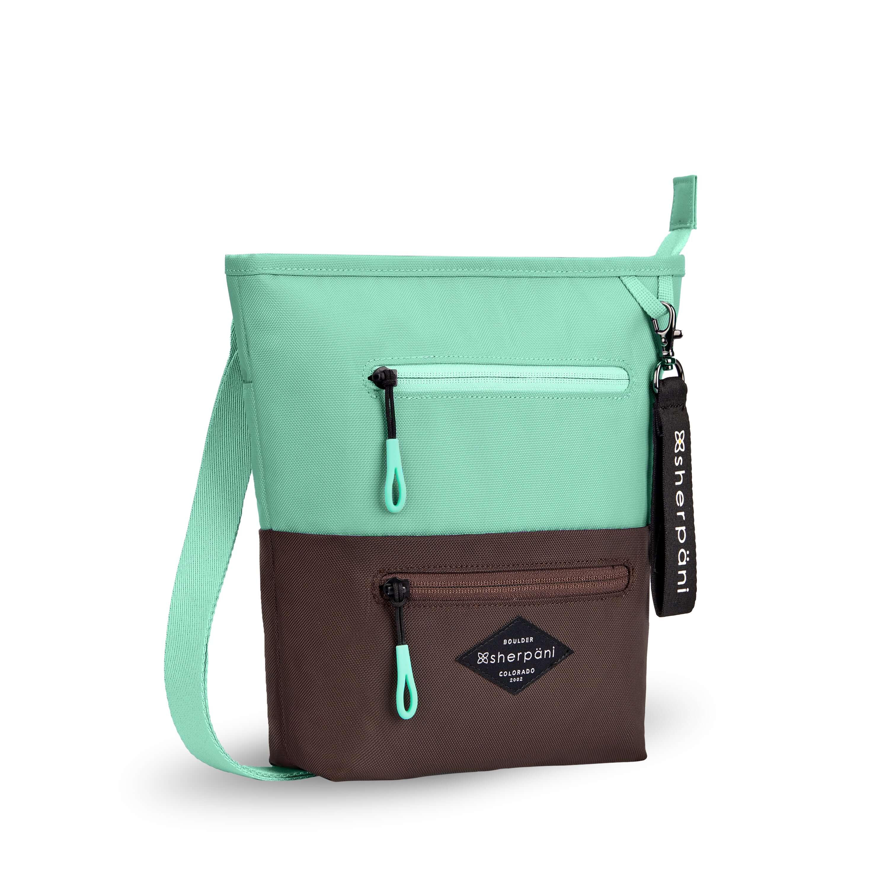 Angled front view of Sherpani’s crossbody, the Sadie, in Seagreen. The top half of the bag is light green and the bottom half of the bag is brown. It features two exterior zipper pockets on the front panel with easy-pull zippers accented in light green. A Sherpani branded keychain is attached to a fabric loop in the upper right corner. It has an adjustable crossbody strap.