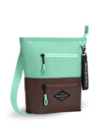 Angled front view of Sherpani’s crossbody, the Sadie, in Seagreen. The top half of the bag is light green and the bottom half of the bag is brown. It features two exterior zipper pockets on the front panel with easy-pull zippers accented in light green. A Sherpani branded keychain is attached to a fabric loop in the upper right corner. It has an adjustable crossbody strap.