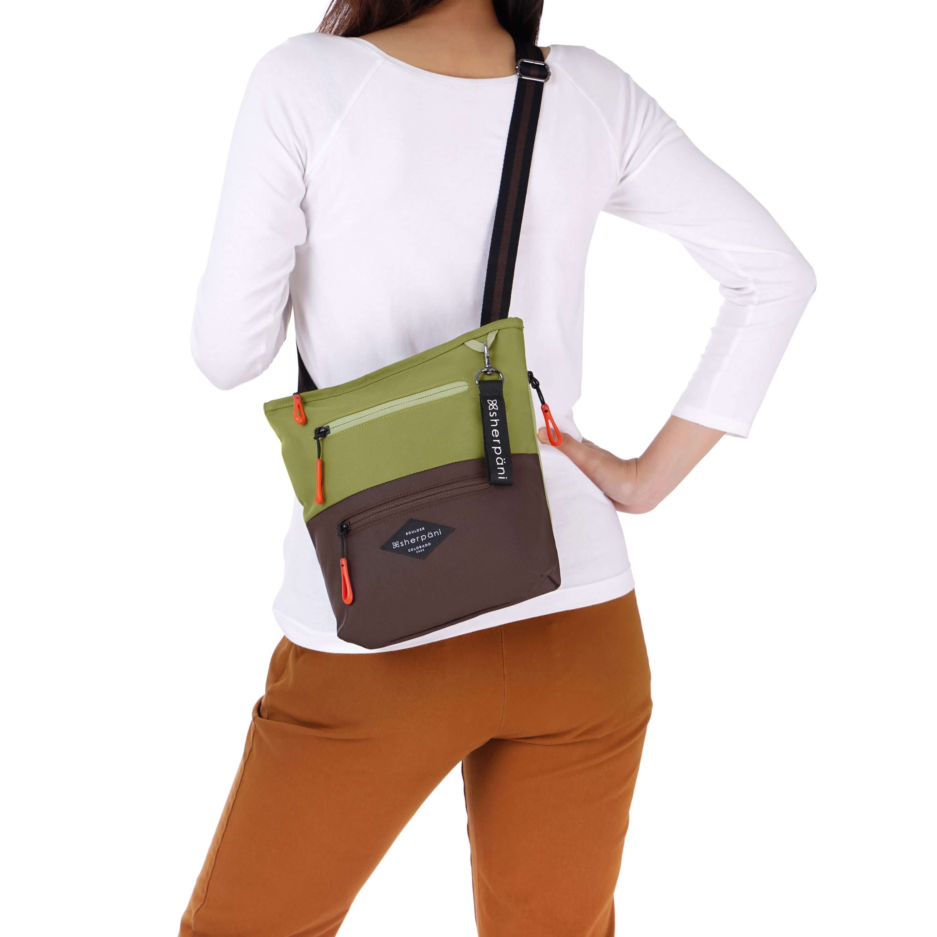 Close up view of a dark haired model facing away from the camera. She is wearing a white shirt and orange pants. She carries Sherpani crossbody, the Sadie in Cactus. 