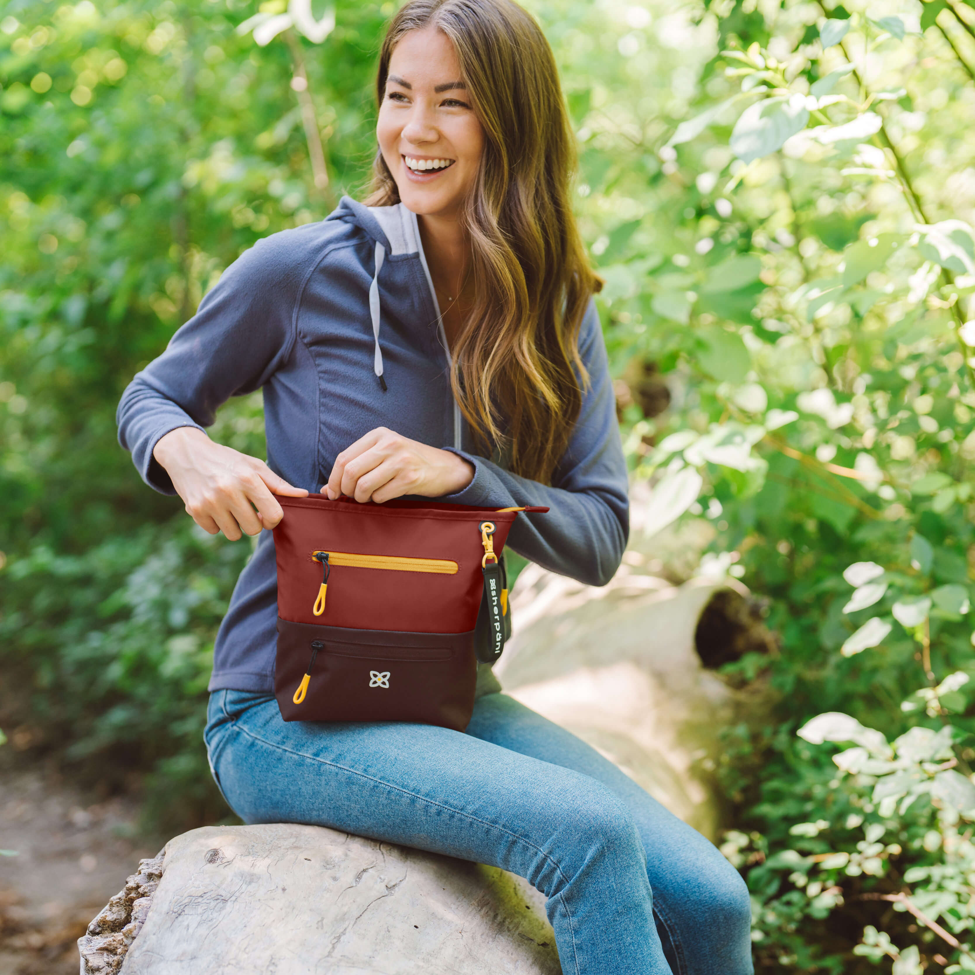 A smiling woman sits outside on a log. She is opening the zipper of her Sherpani travel bag, the Sadie in Cider. 