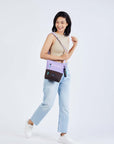 Full body view of a model walking to the side and smiling over her right shoulder. She is wearing a tan tank top, faded jeans and white sneakers. She carries Sherpani's crossbody, the Sadie in Lavender, over her shoulder.
