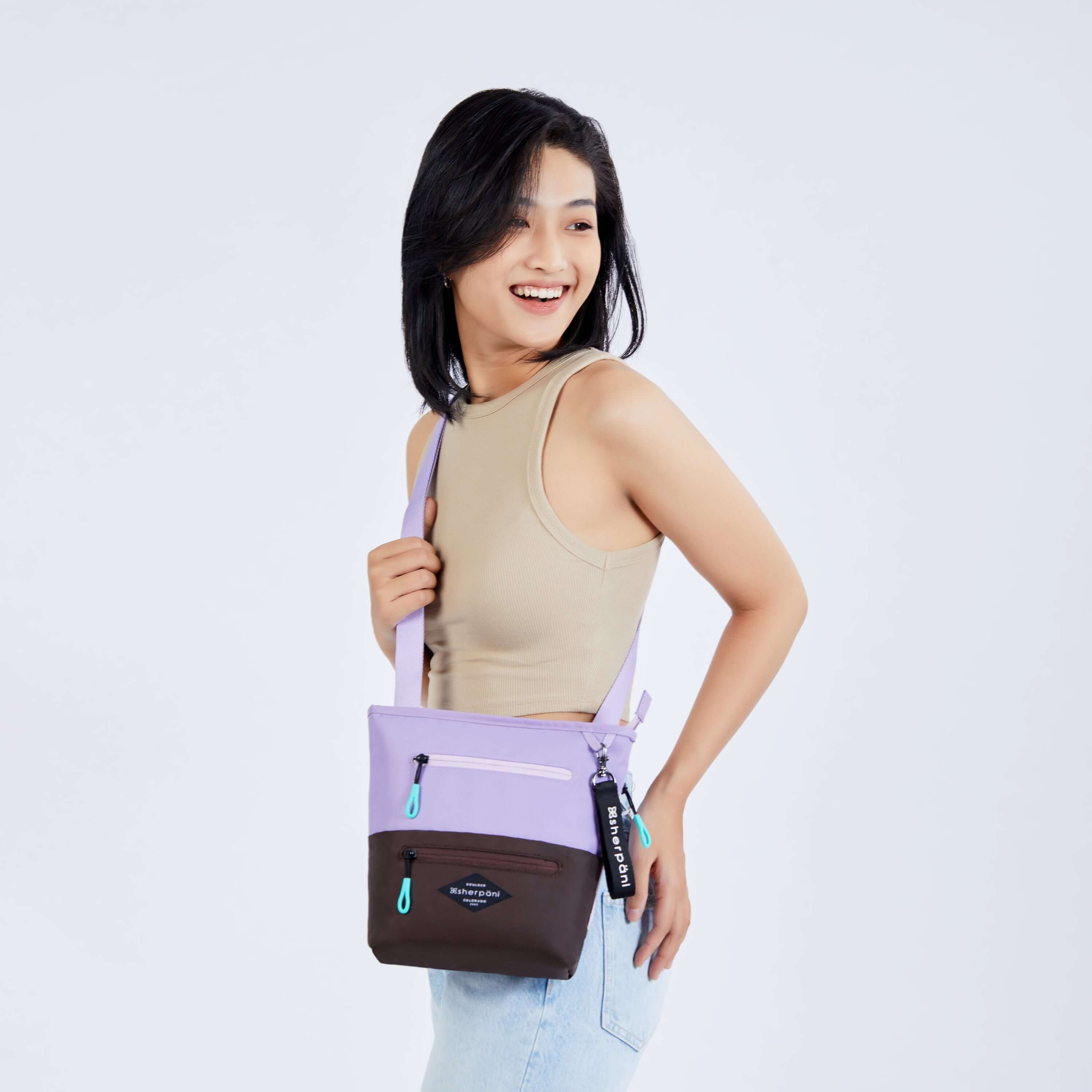 Close up view of a dark haired model facing the side and smiling over her left shoulder. She is wearing a tan tank top and faded jeans. She carries Sherpani&#39;s crossbody, the Sadie in Lavender, as a crossbody.