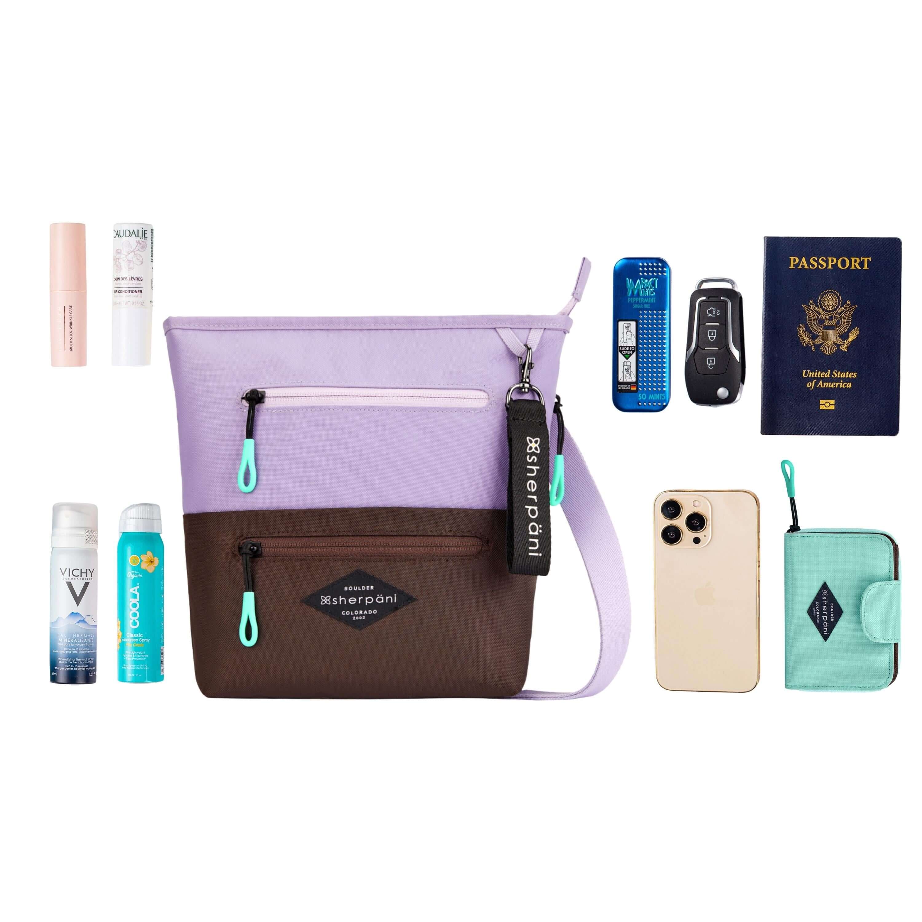 Top view of example items to fill the bag. Sherpani crossbody, the Sadie in Lavender, lies in the center. It is surrounded by an assortment of items: skincare products, sunscreen, body spray, mints, car key, passport, phone and Sherpani travel accessory the Barcelona in Seagreen.