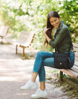 A woman sits on a park bench. She is wearing Sherpani purse with RFID protection, the Sadie in Raven.