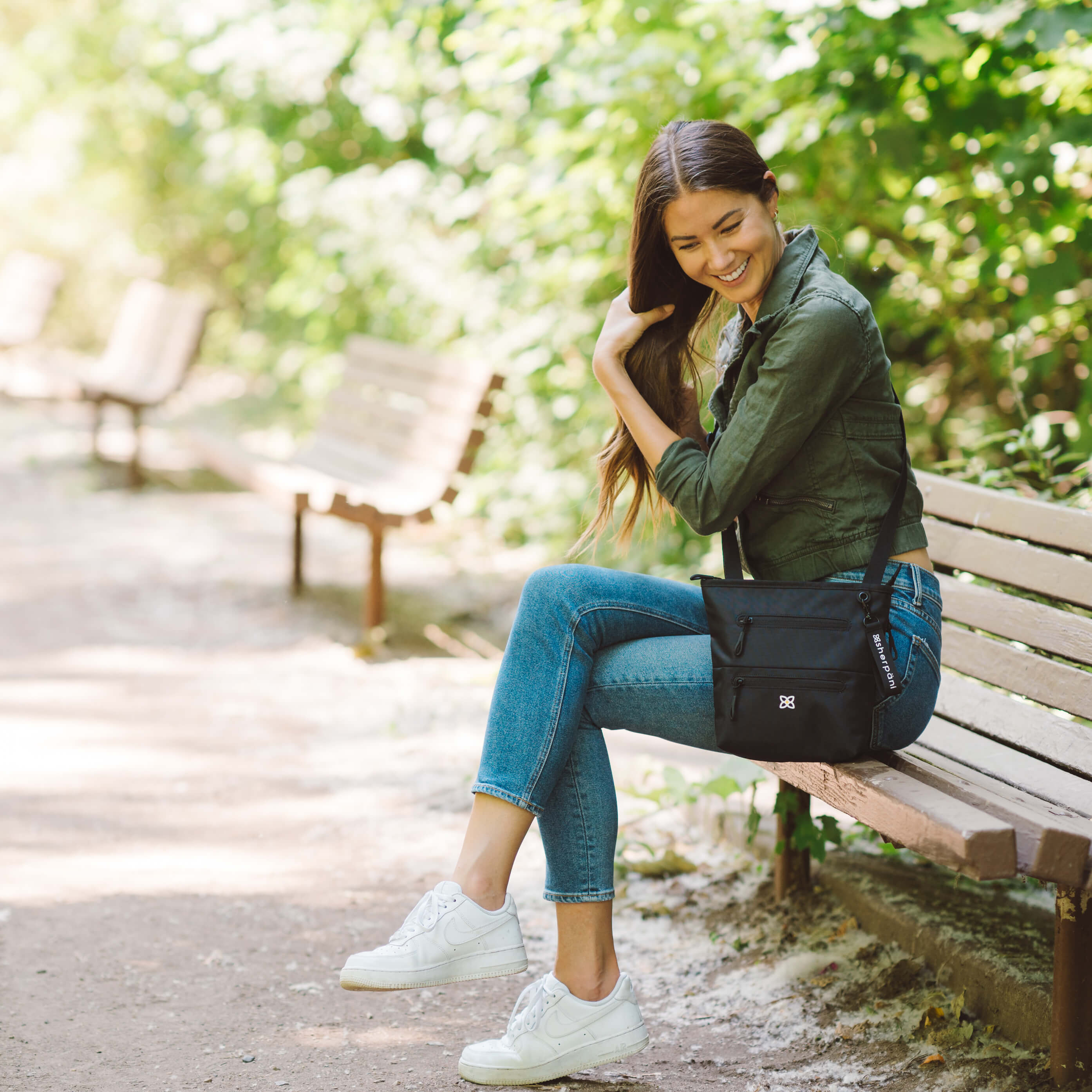 A woman sits on a park bench. She is wearing Sherpani purse with RFID protection, the Sadie in Raven. 