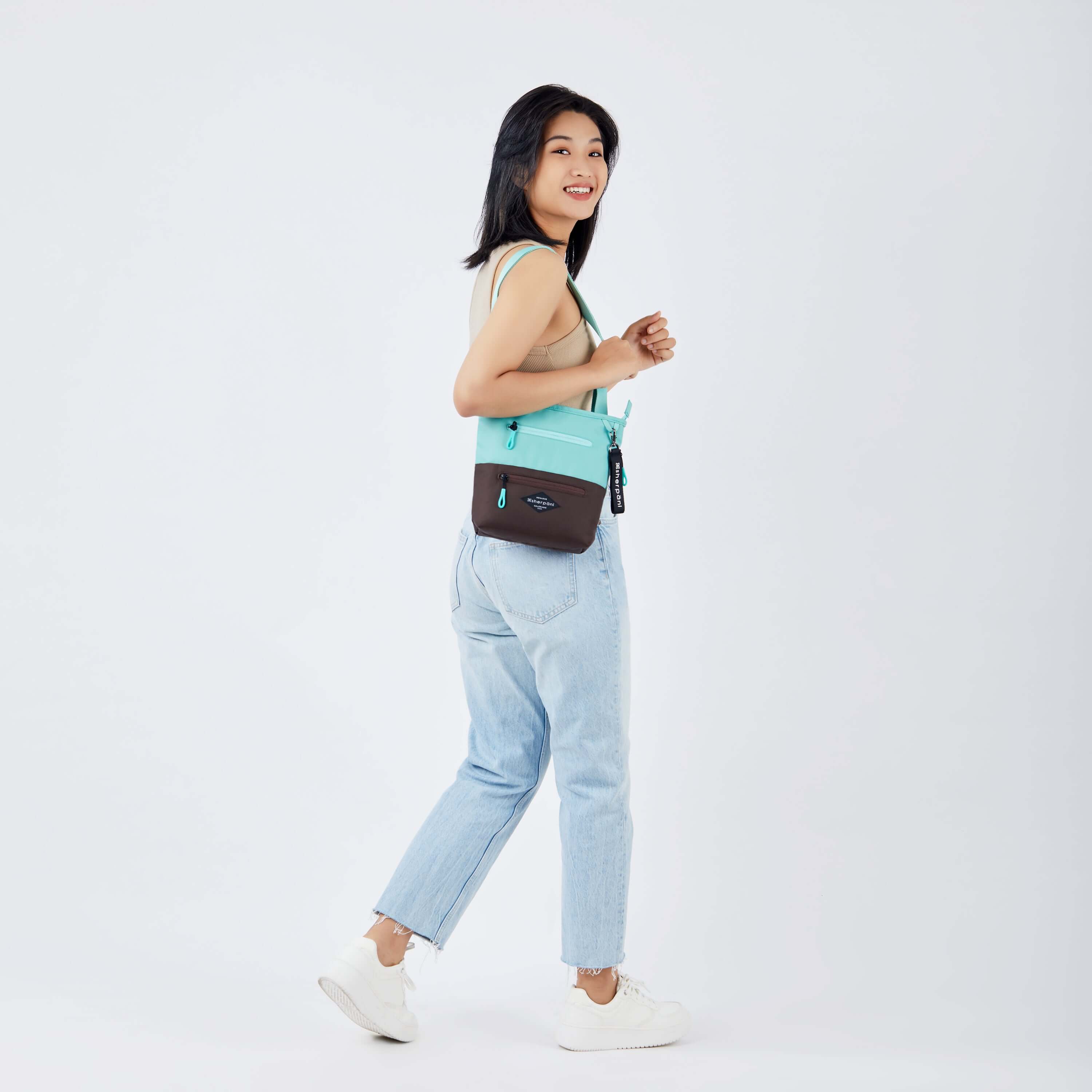 Full body view of a dark haired model facing away from the camera and smiling over her right shoulder. She is wearing a tan tank top, faded jeans and white sneakers. She carries Sherpani's crossbody, the Sadie in Seagreen, over her shoulder. 