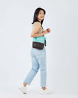 Full body view of a dark haired model facing away from the camera and smiling over her right shoulder. She is wearing a tan tank top, faded jeans and white sneakers. She carries Sherpani's crossbody, the Sadie in Seagreen, over her shoulder.