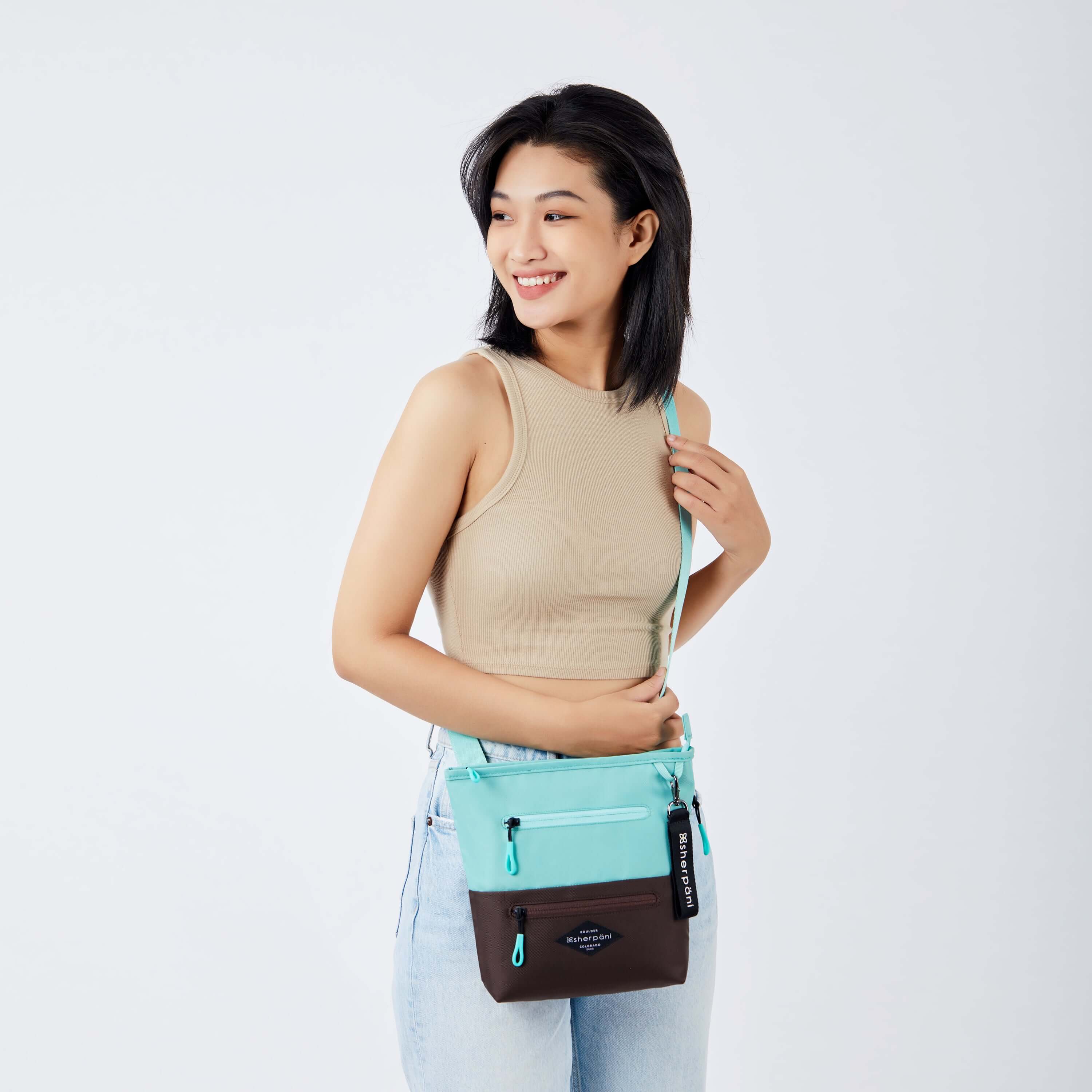 Close up view of a dark haired model smiling over her right shoulder. She is wearing a tan tank top and faded jeans. She carries Sherpani's crossbody, the Sadie in Seagreen, as a crossbody. 