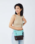 Close up view of a dark haired model smiling over her right shoulder. She is wearing a tan tank top and faded jeans. She carries Sherpani's crossbody, the Sadie in Seagreen, as a crossbody.