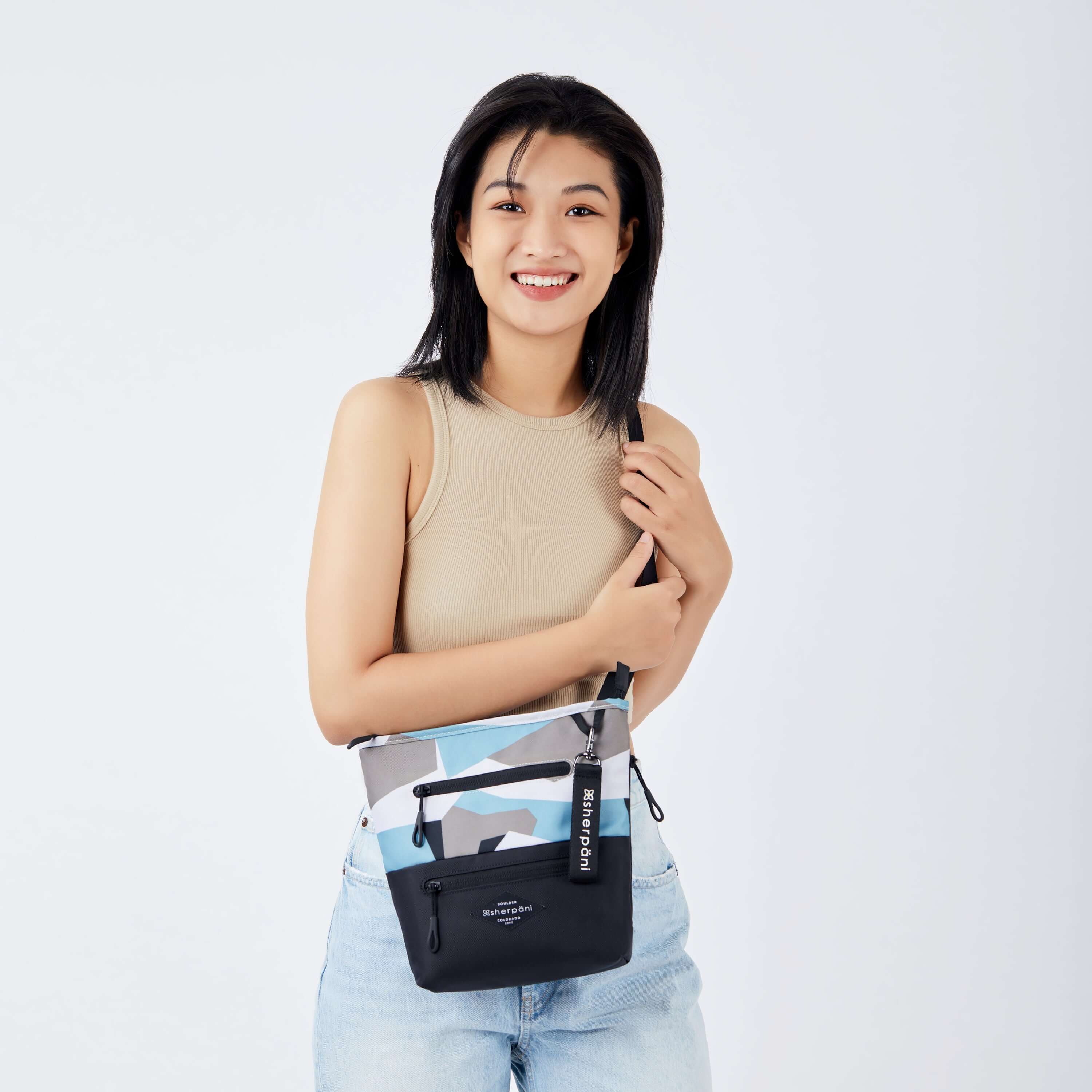Close up view of a dark haired model facing the camera and smiling. She is wearing a tan tank top and faded jeans. She carries Sherpani's crossbody, the Sadie in Summer Camo, as a crossbody. 
