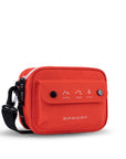 Angled front view of Sherpani crossbody, the Osaka in Poppy. The bag is red in color with a black daisy chain, accent buttons and adjustable/detachable crossbody strap. There is an external pocket on the front of the bag with a fold over flap. On the flap are white graphics of mountains, a wave and a pine tree above white text that reads "Open Your Eyes."