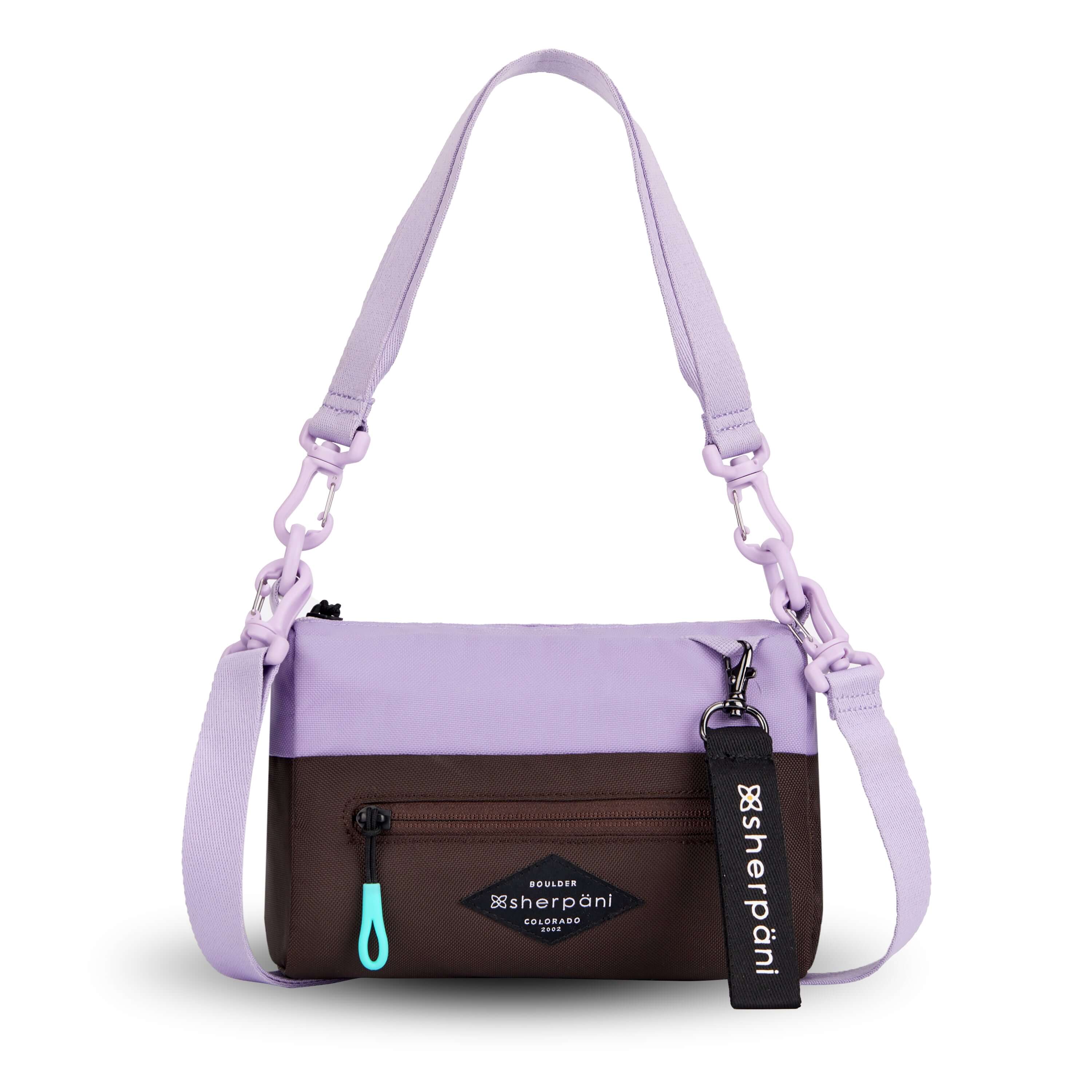 Flat front view of Sherpani's purse, the Skye in Lavender. It is two-toned, the top half of the bag is lavender and the bottom half of the bag is brown. There is an external zipper compartment with an easy-pull zipper accented in aqua. There is a branded Sherpani keychain clipped to the upper right corner. The bag has a detachable short strap and a longer detachable/adjustable crossbody strap. 