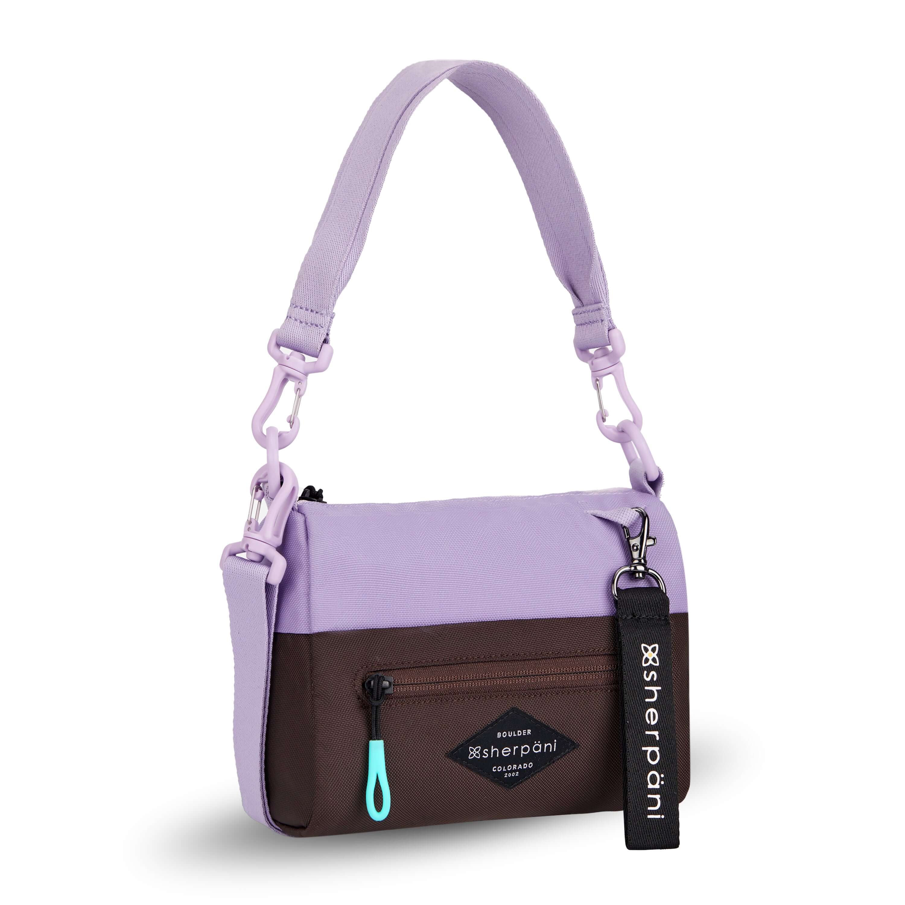 Angled front view of Sherpani's purse, the Skye in Lavender. It is two-toned, the top half of the bag is lavender and the bottom half of the bag is brown. There is an external zipper compartment with an easy-pull zipper accented in aqua. There is a branded Sherpani keychain clipped to the upper right corner. The bag has a detachable short strap and a longer detachable/adjustable crossbody strap. 