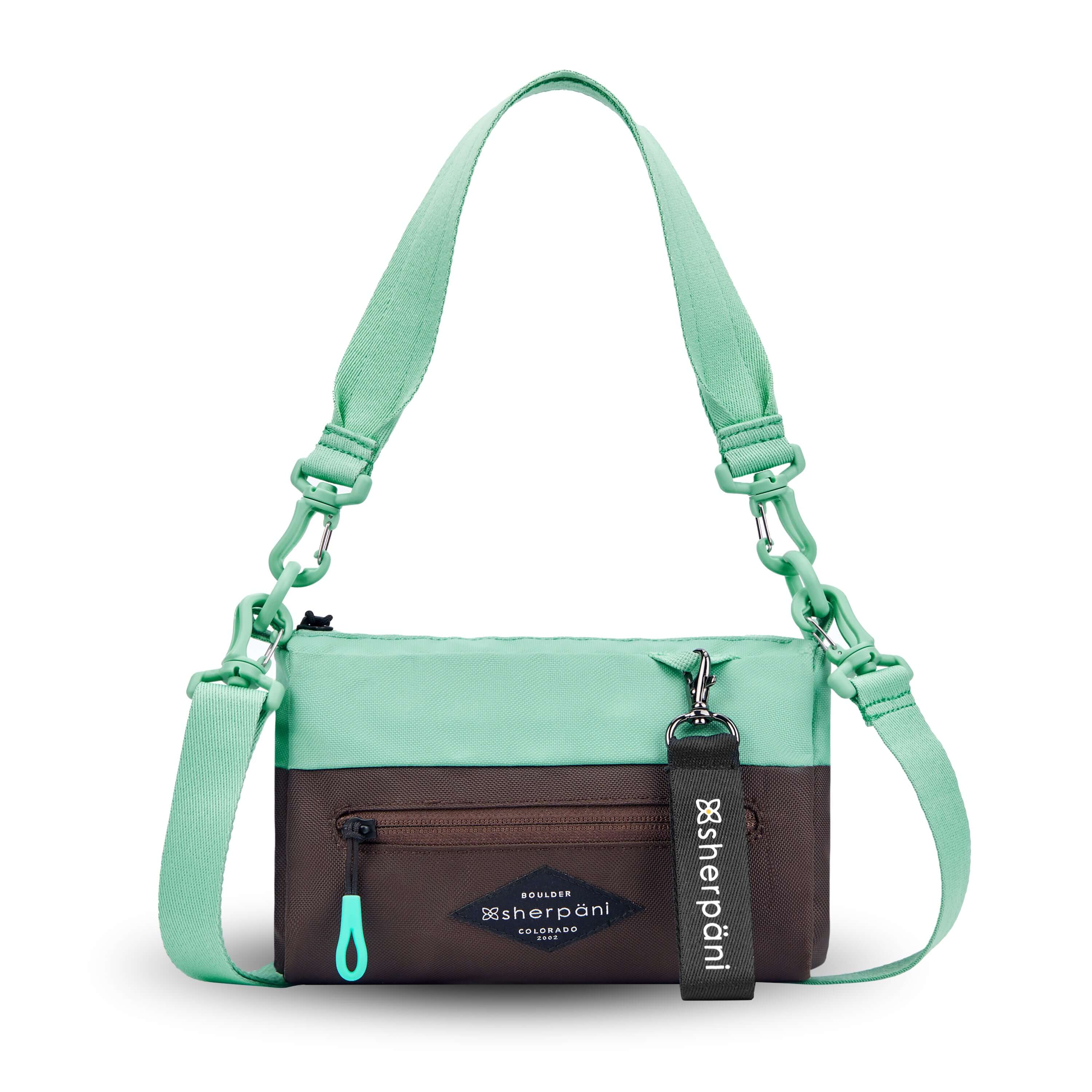 Flat front view of Sherpani's purse, the Skye in Seagreen. It is two-toned, the top half of the bag is light green and the bottom half of the bag is brown. There is an external zipper compartment with an easy-pull zipper accented in light green. There is a branded Sherpani keychain clipped to the upper right corner. The bag has a detachable short strap and a longer detachable/adjustable crossbody strap. 