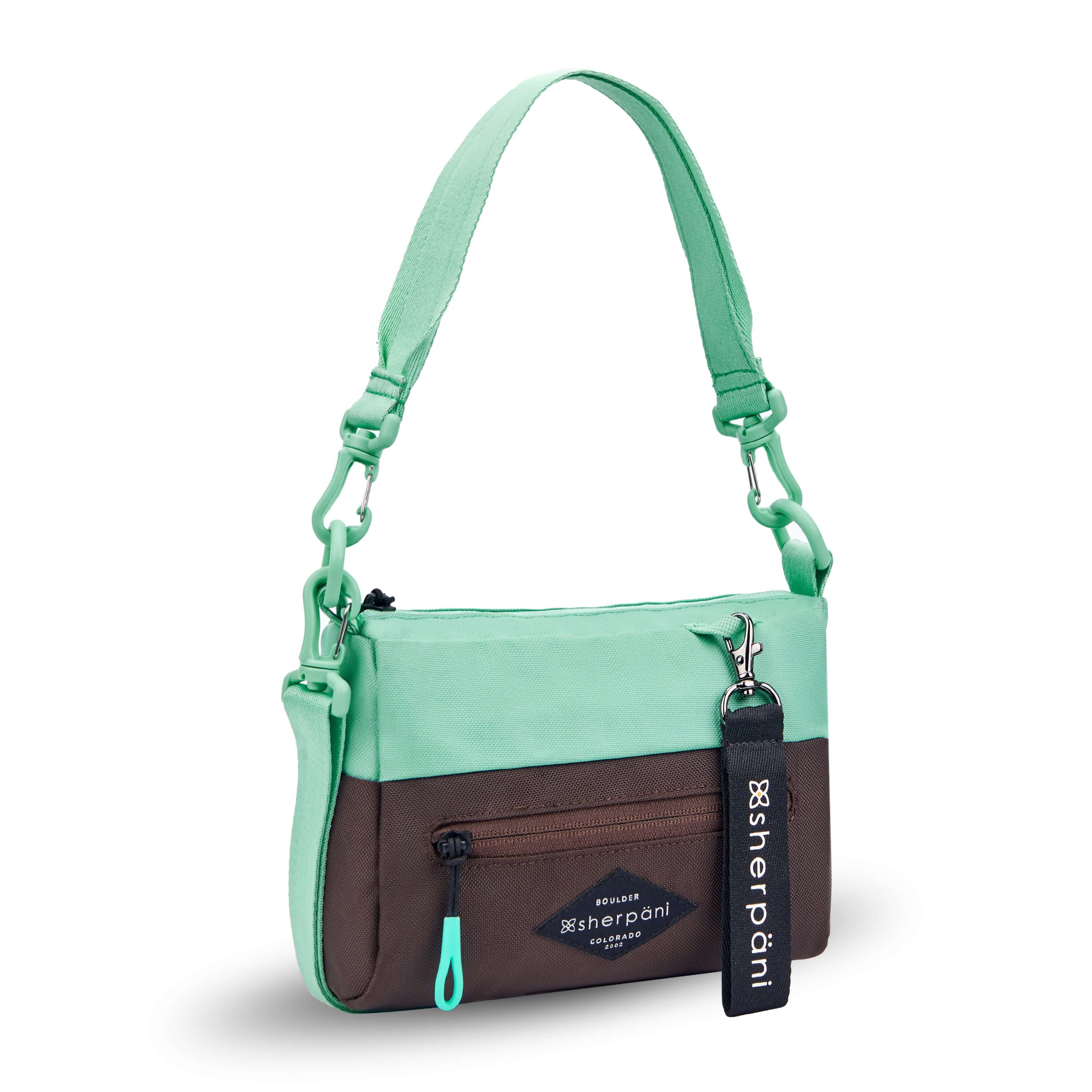Angled front view of Sherpani's purse, the Skye in Seagreen. It is two-toned, the top half of the bag is light green and the bottom half of the bag is brown. There is an external zipper compartment with an easy-pull zipper accented in light green. There is a branded Sherpani keychain clipped to the upper right corner. The bag has a detachable short strap and a longer detachable/adjustable crossbody strap. #color_seagreen