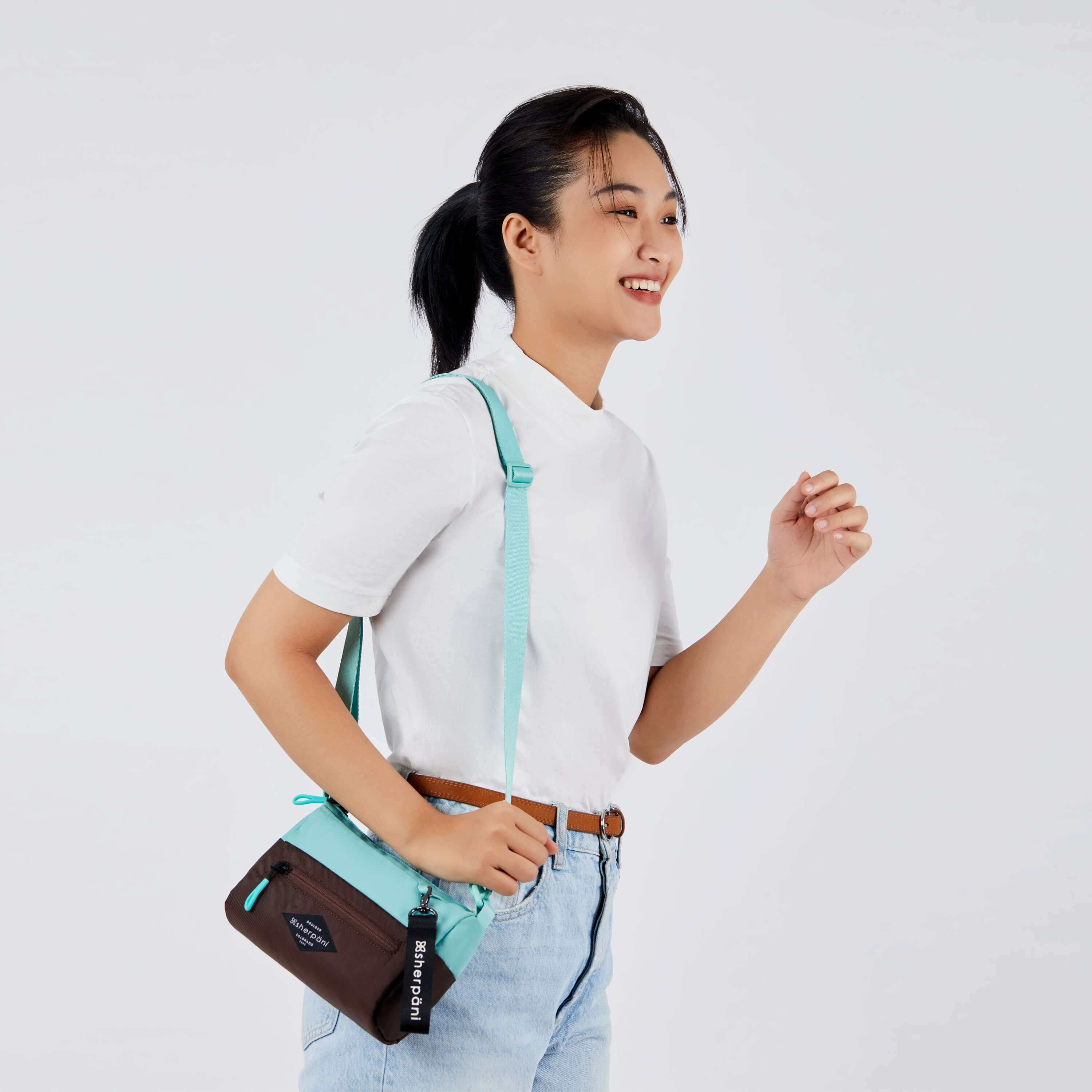 Close up view of a dark haired model facing the side and smiling. She is wearing a white tee shirt and jeans. She carries Sherpani's purse, the Skye in Seagreen, over her shoulder. 