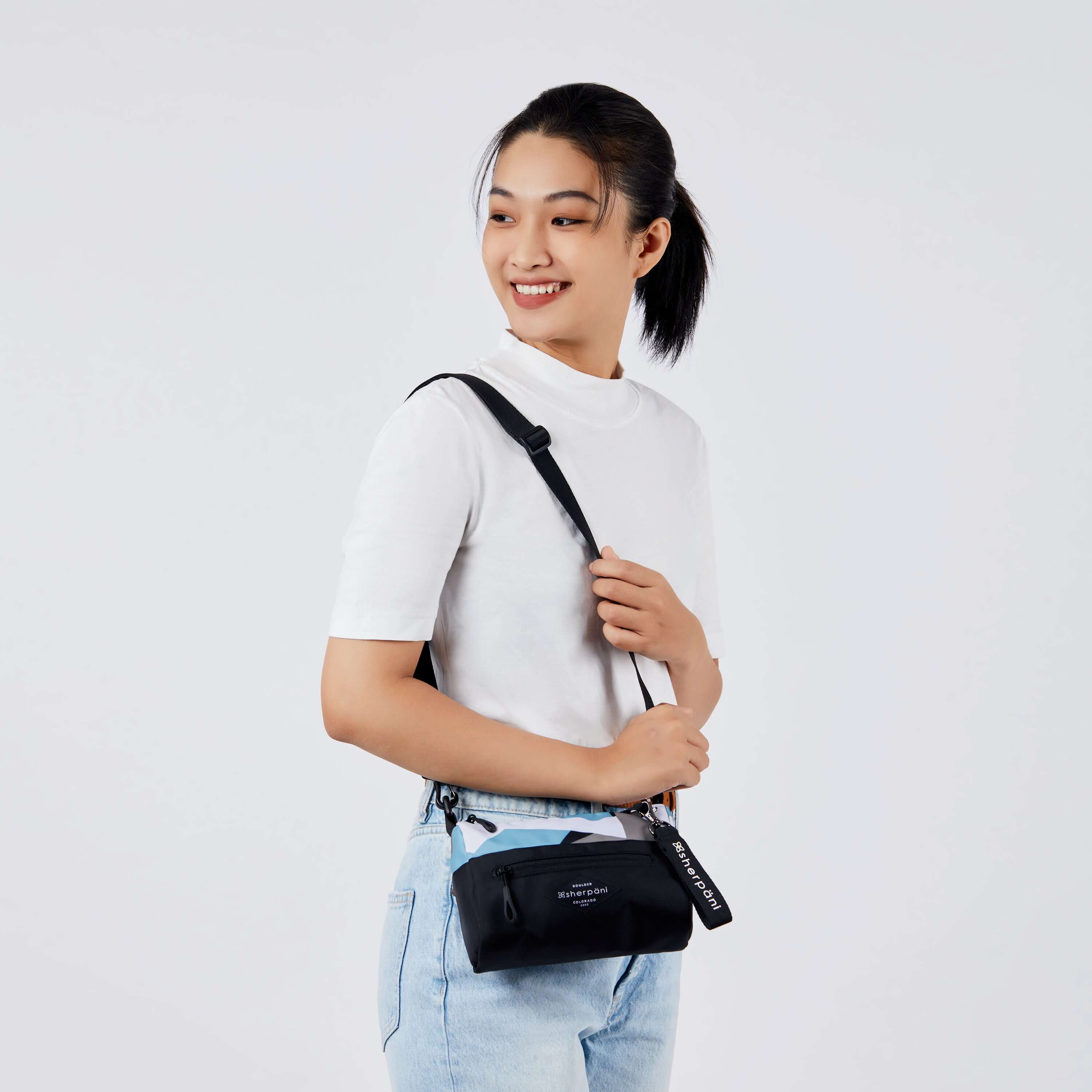 Close up view of a dark haired model facing the side and smiling over her right shoulder. She is wearing a white tee shirt and jeans. She carries Sherpani's purse, the Skye in Summer Camo, over her shoulder. 