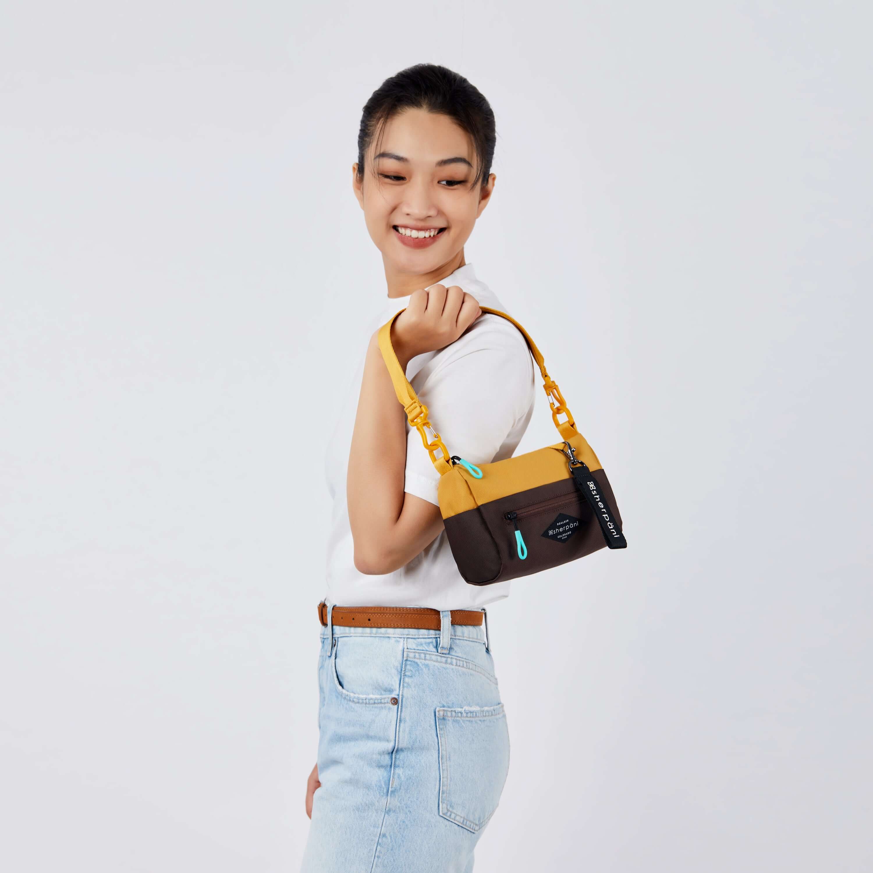 Close up view of a dark haired model facing the side and smiling over her left shoulder. She is wearing a white tee shirt and jeans. She carries Sherpani's purse, the Skye in Sundial, over her shoulder. 