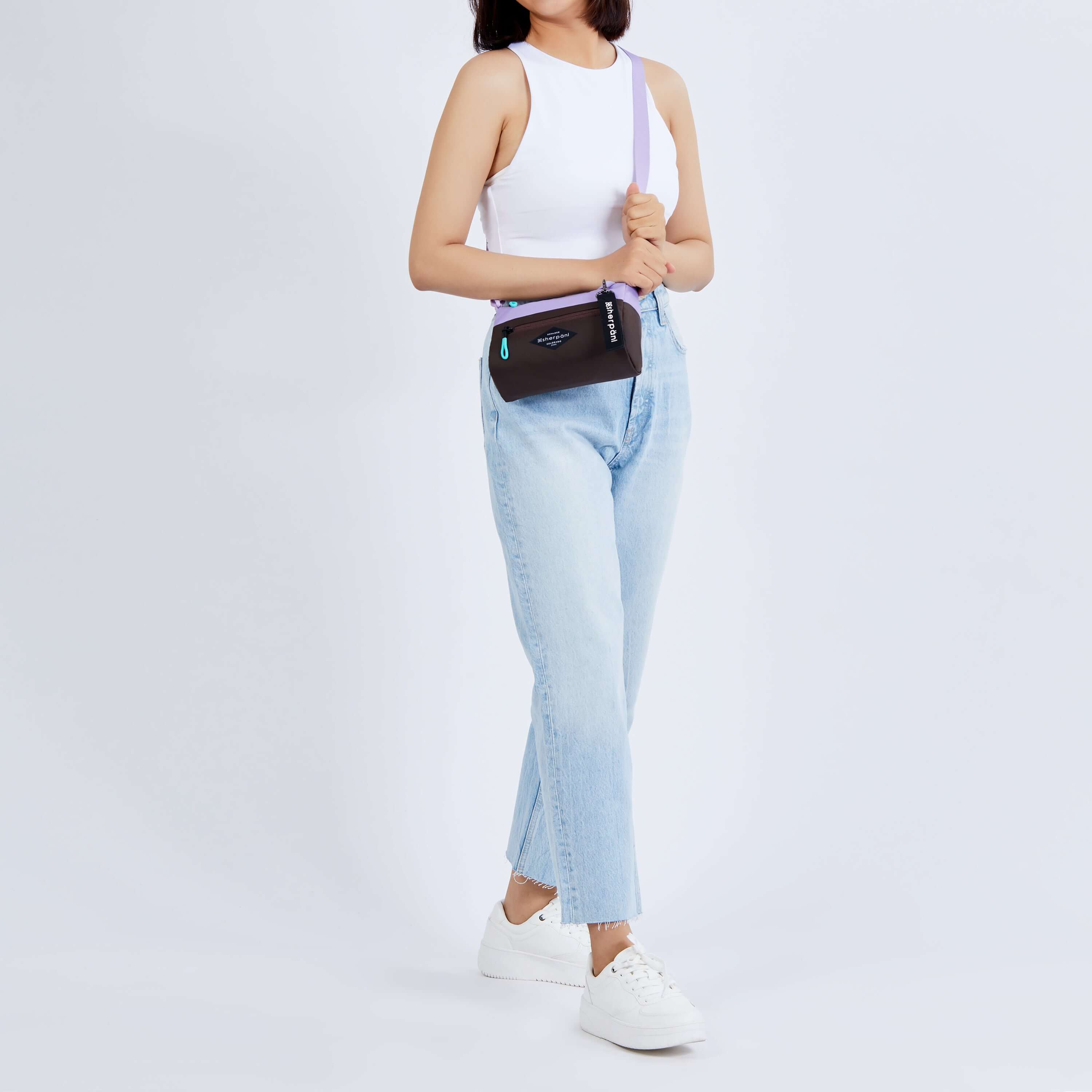 Full body view of a dark haired model facing the camera. She is wearing a white tank top, jeans and white sneakers. She carries Sherpani's purse, the Skye in Lavender, as a crossbody. 