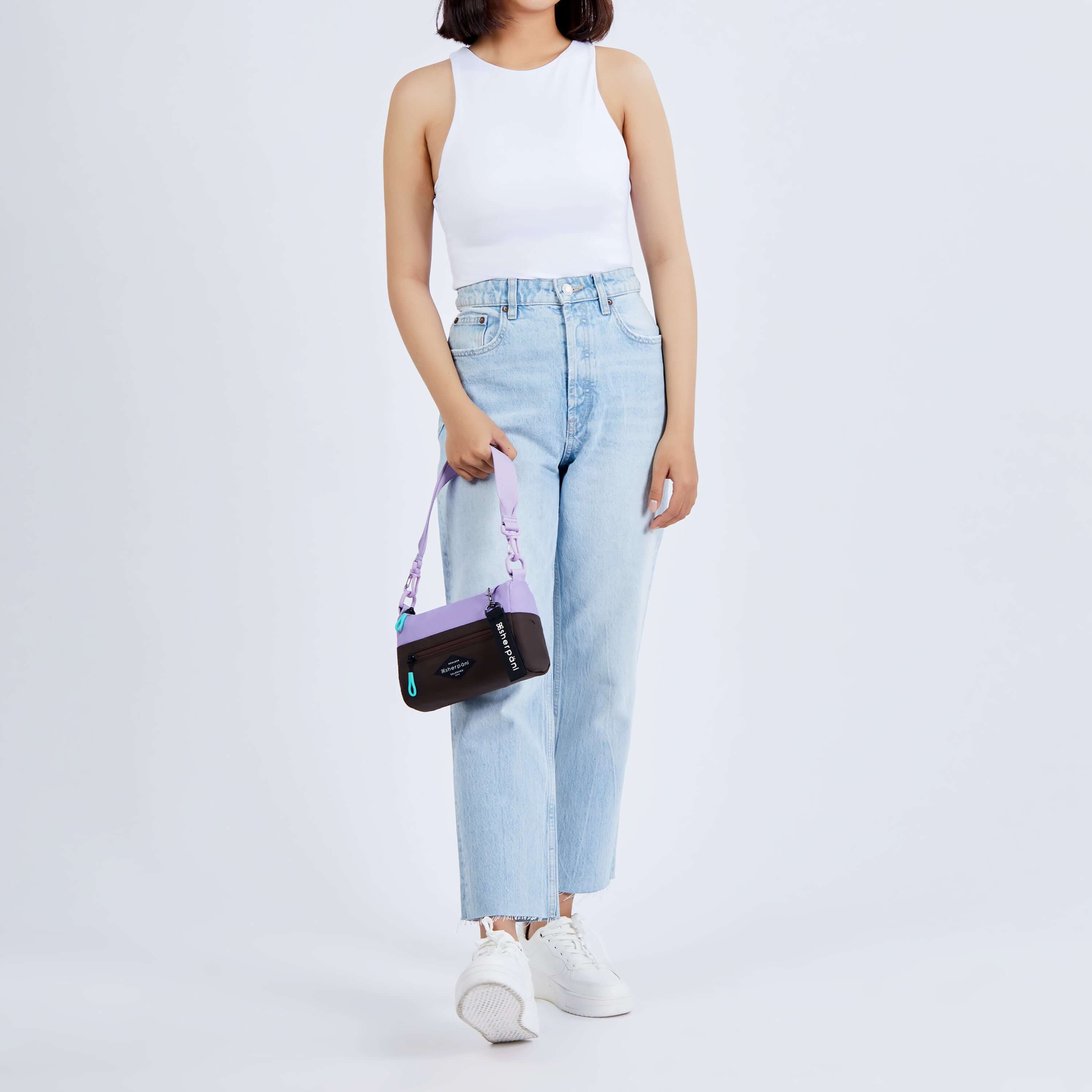Full body view of a dark haired model facing the camera. She is wearing a white tank top, jeans and white sneakers. She carries Sherpani's purse, the Skye in Lavender, by her side. 